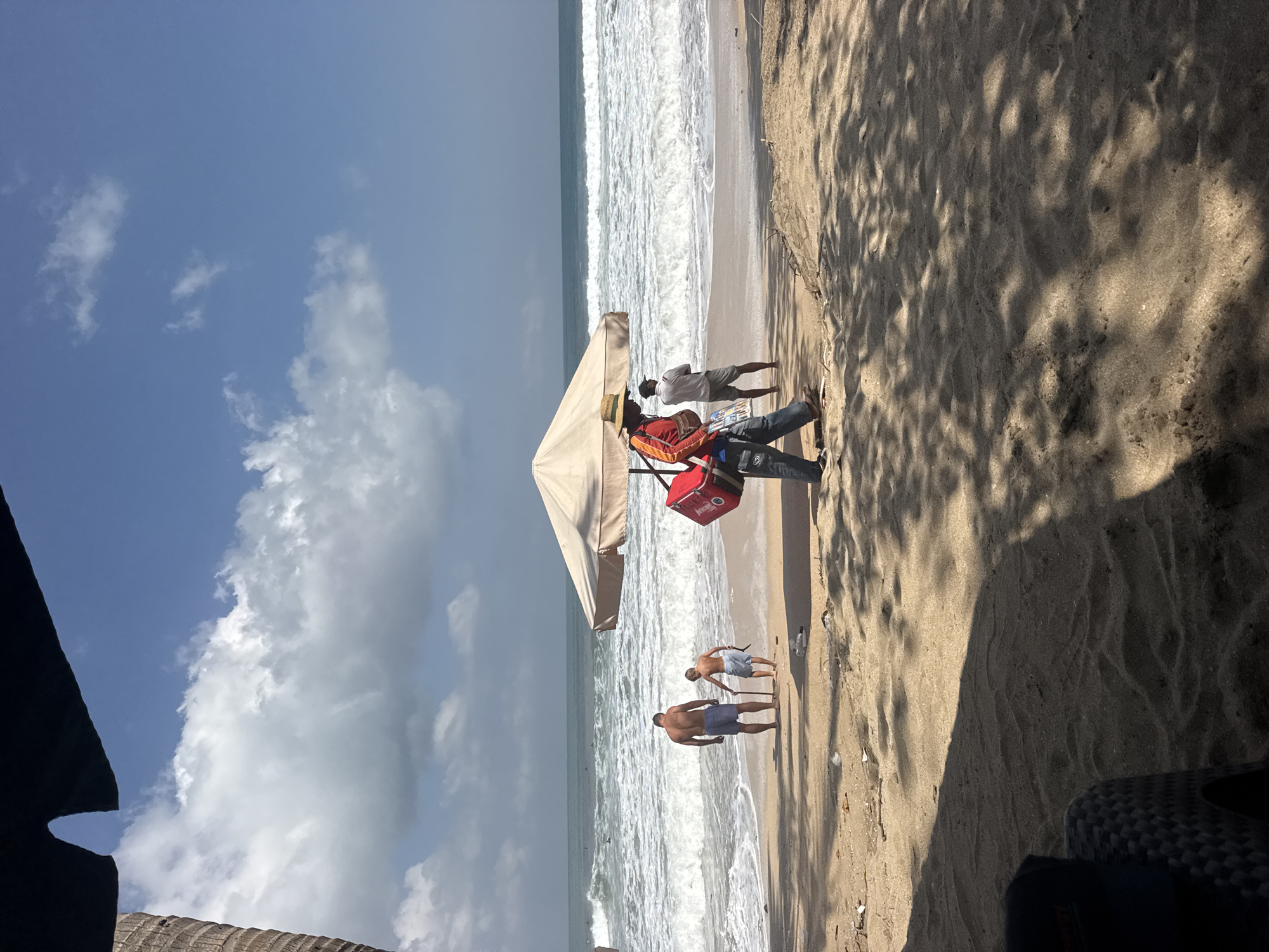 The image shows a beach scene in Kuta, Indonesia. A man and a child are standing on the sand near the water's edge, with waves in the background. A vendor with a red bag and straw hat is walking on the beach under a large umbrella. Another man is standing near the umbrella, looking out at the ocean. The sand is littered with some debris. Shadows from palm trees are cast across the sand. The sky is blue with some white clouds.