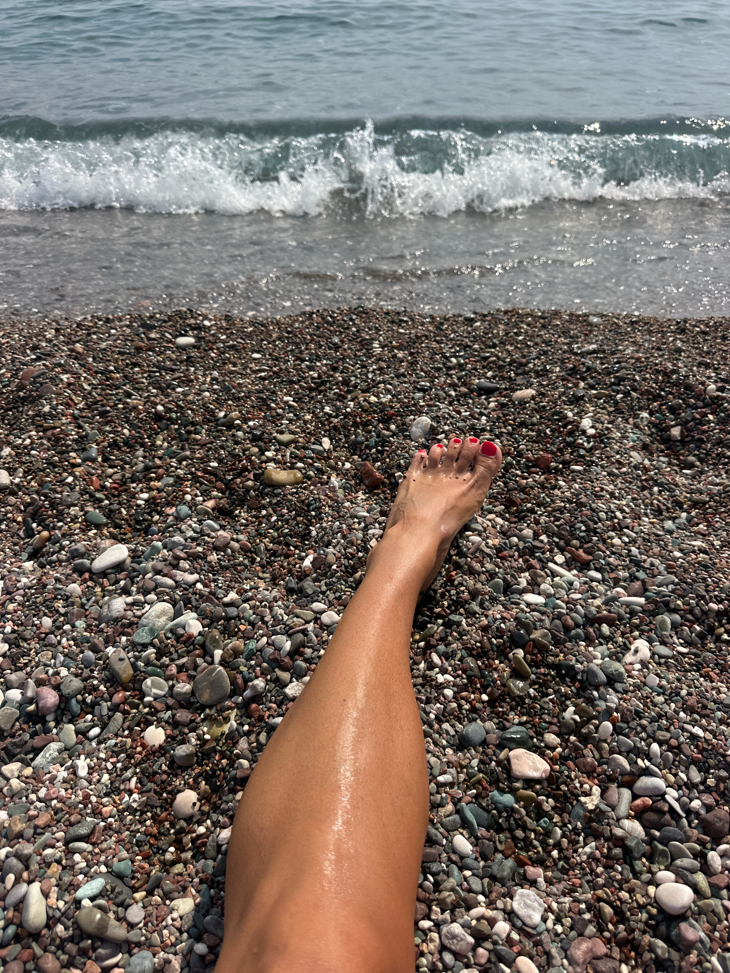 A person's bare leg and foot with red nail polish are positioned on a pebble beach. The leg is wet and glistening. In the background, the calm blue sea laps against the shore, with gentle waves breaking. The pebbles on the beach are small and varied in color, forming a textured ground. The scene is set on a beach in Boreti, Montenegro.