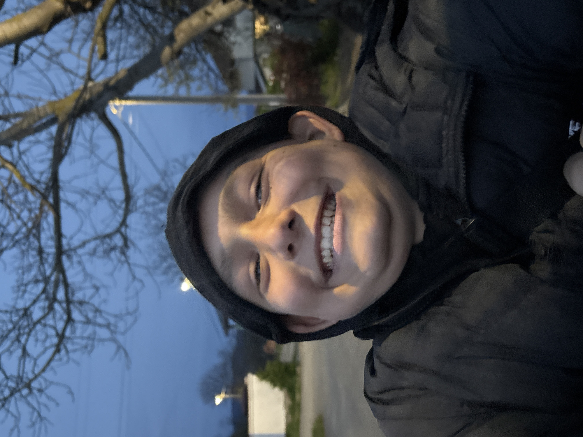 A smiling young individual is centered in the image, wearing a dark hooded jacket. The person's face is well-lit, showing teeth in a wide smile. Behind the person, a street scene is visible, including bare trees and what appears to be a street light. The setting appears to be outdoors, possibly on a street or sidewalk in the evening or early morning.