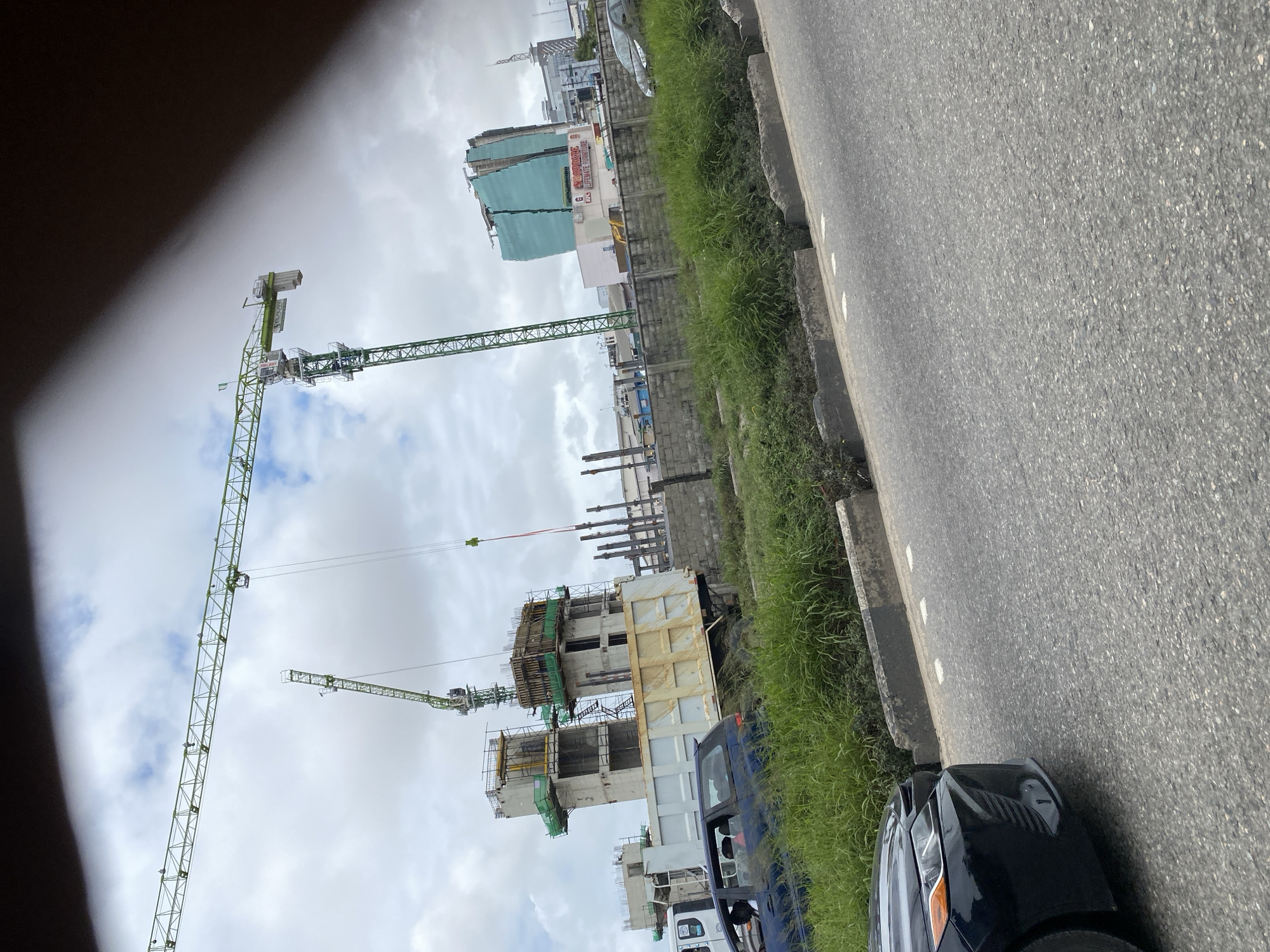 In Lekki Phase I, Nigeria, a construction site is visible with multiple cranes, including two prominent green tower cranes. One crane's jib extends horizontally across the frame, while the other is angled upwards. In the background, a partially constructed building stands, with concrete pillars and formwork evident. Adjacent to it, a taller, modern building with a teal-colored exterior is under construction, wrapped in netting. Several vehicles are present, including a large dump truck, a dark blue car in the foreground, and other cars further down the road. The scene is bordered by a concrete wall and lush green vegetation. The sky is overcast with clouds.