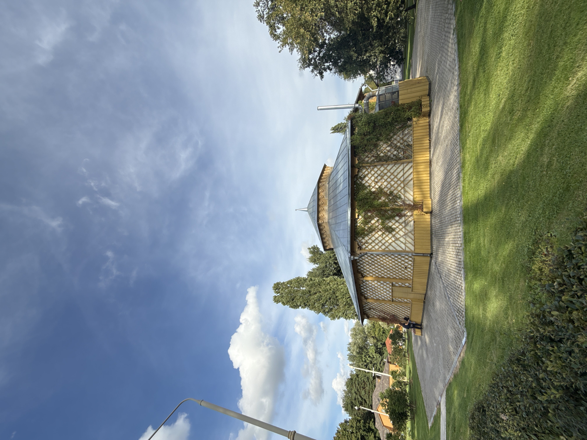 A wide, low-angle shot captures a circular wooden pavilion with a metal roof in a park-like setting. The pavilion has a yellow base with a lattice design on its walls, partially covered by climbing ivy. A pointed finial adorns the roof's peak. To the left, a tall, gray lamppost stands against a bright blue sky with scattered white clouds. In the background, green trees and several houses with orange and red roofs are visible. A paved walkway surrounds the pavilion, and a section of neatly trimmed green grass occupies the foreground. A lone person stands near the pavilion entrance, facing away from the camera and interacting with something on the ground. The scene suggests a sunny day in an outdoor public space, likely a park or botanical garden, possibly within the city of Moscow, Russia.