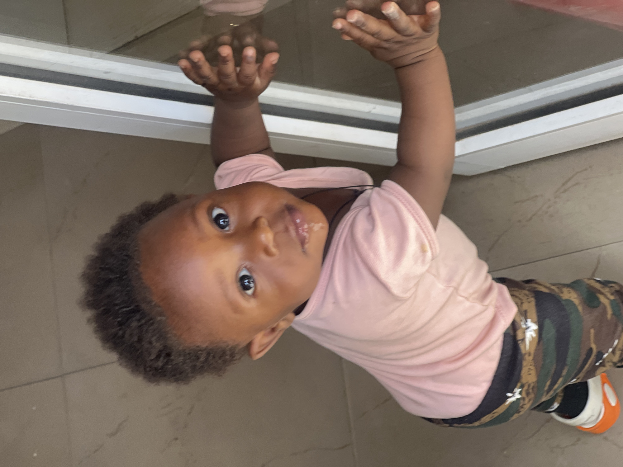 A toddler with brown skin, short curly hair, and wearing a pink short-sleeve shirt and camouflage pants is holding onto a window frame with both hands. The toddler is looking directly at the camera. The location appears to be indoors on a tile floor. The specific location in the city of Gura Top, Nigeria cannot be determined from this image.