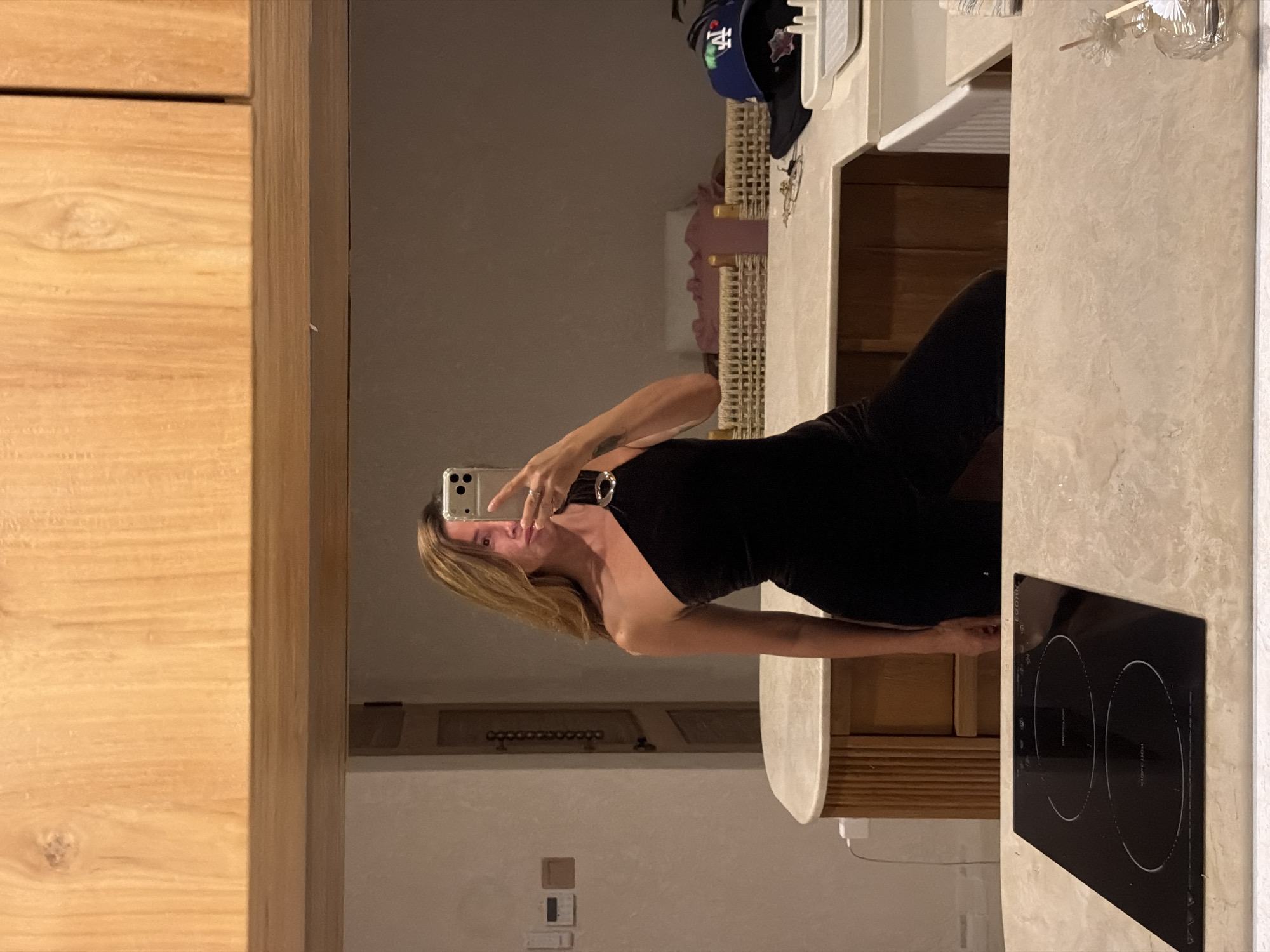 The image shows a woman taking a mirror selfie in what appears to be a modern kitchen or dining area. She is wearing a black one-shoulder dress with a gold accent at the shoulder. The background includes wooden cabinetry, a countertop, a sink, an induction cooktop, and some stored items like a hat. The setting is likely indoors, possibly in a residential or hotel space. The location specified, Mengwi, Indonesia, does not appear to be directly evident from visual cues in the image.