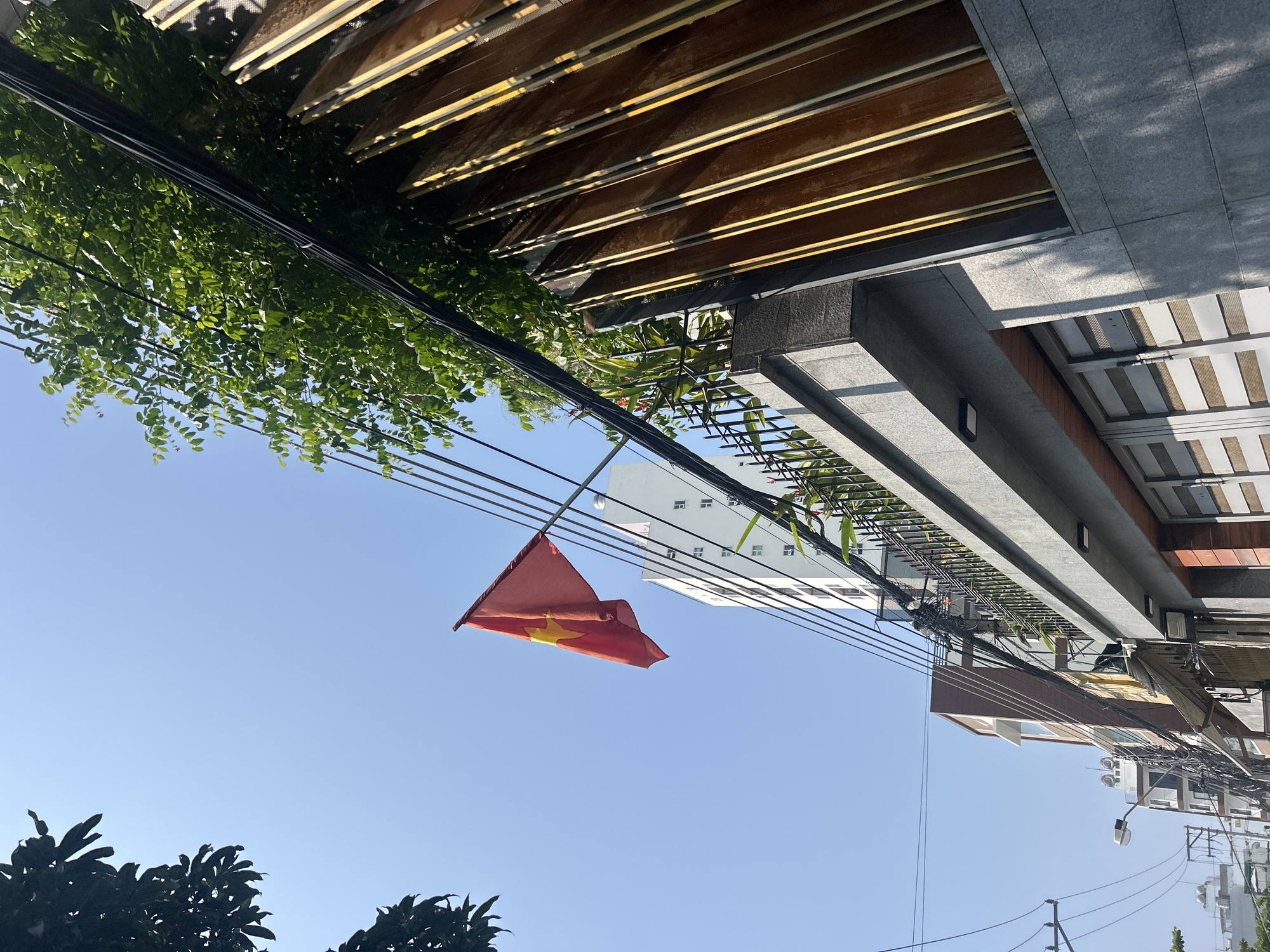 The image, taken in Bắc Nha Trang, Vietnam, shows a low-angle view of a street scene under a clear blue sky. A Vietnamese flag hangs suspended from electrical wires that run across the frame. Dense green foliage obscures part of the sky and building structures at the top left. The upper portion of buildings with various architectural features, including exposed brown and grey awnings, stone or concrete ledges, and a white multi-story building in the background, are visible. Electrical wires are prominent throughout the upper half of the image. Trees are visible at the bottom left. A street light is also visible in the lower-right corner.
