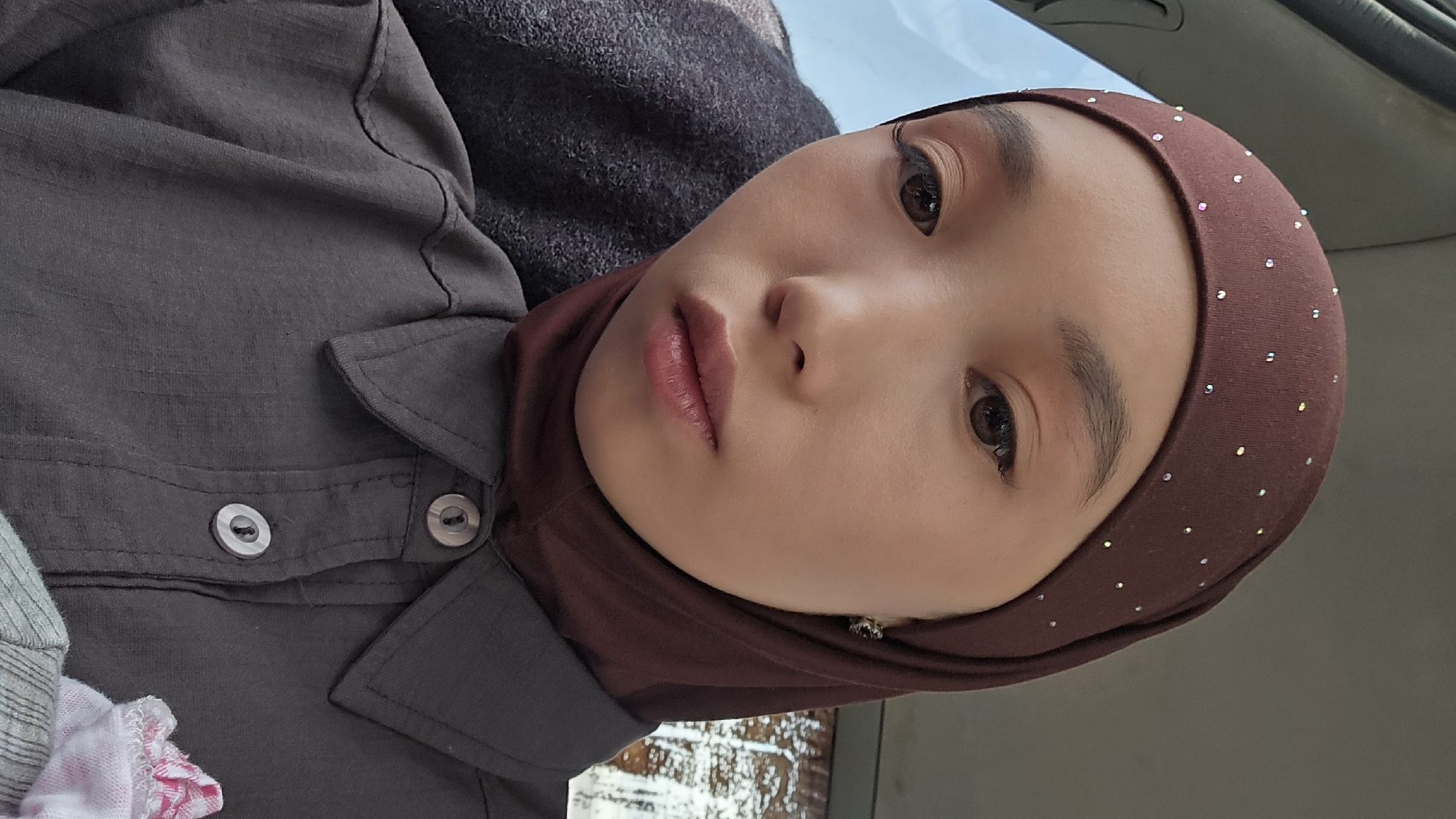 A close-up, selfie-style photograph shows a young woman wearing a brown hijab adorned with small, scattered rhinestones. She is also wearing a dark gray collared shirt with two visible buttons. The woman has fair skin, dark eyes, and is wearing makeup, including eyeliner and eyeshadow. Her lips are slightly parted, revealing a subtle pink lipstick. She is looking directly at the camera with a neutral expression. The background appears to be the interior of a vehicle, with a blurred gray seat visible to her left and a window with a bright, out-of-focus view to her left. The overall lighting suggests it is daytime. The woman is likely within the city of Kyzyl-Suu, Kyrgyzstan, given the context.