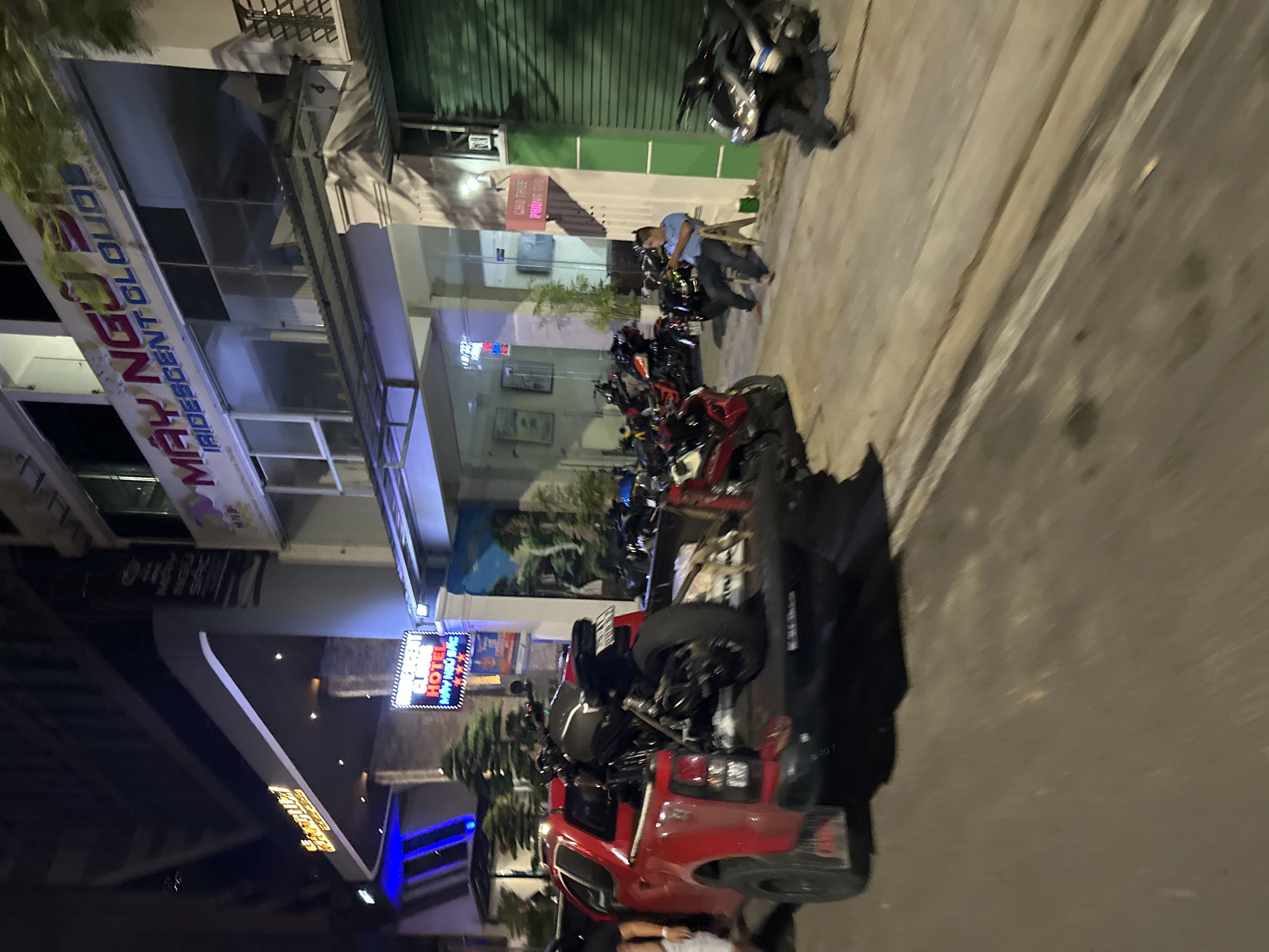 The image depicts a street scene at night in Bac Nha Trang Ward, Vietnam. Parked motorcycles and a red pickup truck dominate the foreground. Several people are present, although their actions and interactions are unclear. Streetlights illuminate buildings with signage in Vietnamese. One sign reads "MAY NGỌC, IRIDESCENT CLOUD". Another sign appears to be a hotel advertisement. The environment consists of buildings, parked vehicles, and people on a city street at night.