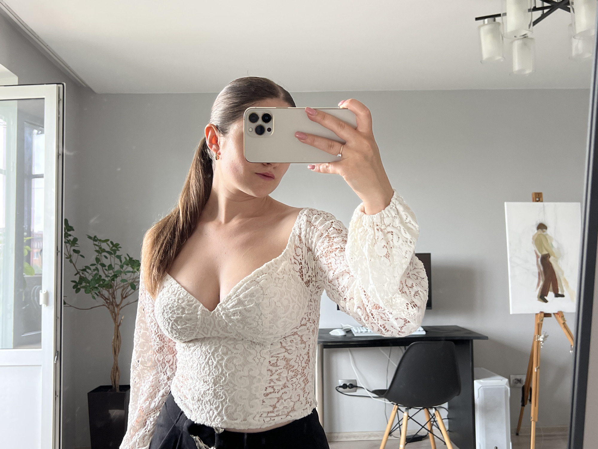 A woman is taking a mirror selfie indoors. She has long brown hair styled in a ponytail and is wearing a white, long-sleeved lace top with a deep V-neck. Her face is obscured by a light-colored smartphone she holds, which features a triple camera array. A ring is visible on her left hand, and she has light pink nail polish. A small hoop earring is visible in her right ear. She is also wearing dark pants or a skirt.
The setting appears to be a room or apartment with light gray walls and a white ceiling. To the left, a white-framed window or door reveals some greenery and a distant building outside, indicating it is daytime. A potted plant with green leaves in a black rectangular pot stands near the window. To the right, a dark desk holds a computer mouse and keyboard, with power cables visible near a wall outlet. A modern black chair with wooden legs is positioned at the desk. Next to the desk, a white air purifier or heater is visible. Further to the right, a wooden easel holds a painting that depicts a standing figure in an abstract or impressionistic style, dressed in light and dark clothing. A chandelier with a black frame and multiple white cylindrical lights hangs from the ceiling, reflected in the mirror. No visible text is present in the image. The location context provided is Ternopil, Ukraine.