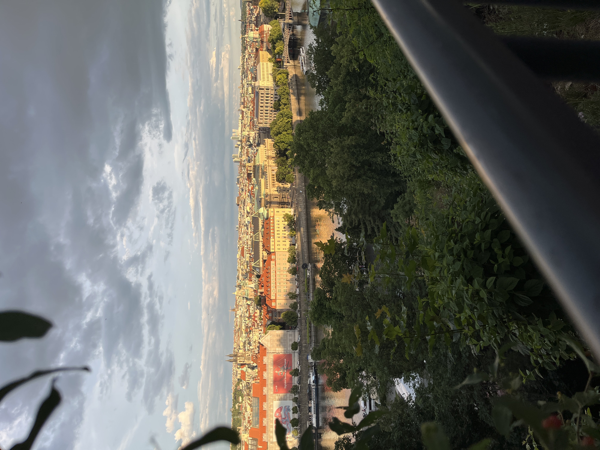 This media file presents an elevated, panoramic view of a European cityscape along a river, bathed in the warm, soft light of late afternoon or early evening. The scene encompasses a wide horizon, featuring numerous buildings, a winding river, and a dynamic sky with clouds.
In the immediate foreground, dark foliage and a metallic railing partially obscure the bottom and right edges, suggesting the photograph was taken from a viewing point on a hill or balcony. Beyond this, the river, likely the Vltava, flows through the city, reflecting the sky and buildings. Several boats, including larger tour vessels and smaller craft, are visible on the water. Bridges span the river at various points.
The city itself is a dense collection of buildings, showcasing a mix of historical and more modern architecture. Many structures have traditional red-tiled roofs, while others feature domes and spires characteristic of Central European cities, strongly resembling Prague. A notable feature is a large building by the river with a prominent red banner on its facade. Partially legible text on this banner includes "digitalni budoucnost" and what appears to be "Moneta Money Bank." Roads line the riverbanks, with cars visible, indicating ongoing urban activity.
The sky above is a blend of light and dark clouds, with patches of blue and areas illuminated by the setting sun, casting a golden glow on some of the buildings in the distance. The overall impression is one of a vibrant, historic city unfolding along its natural waterway. There are no visible people.