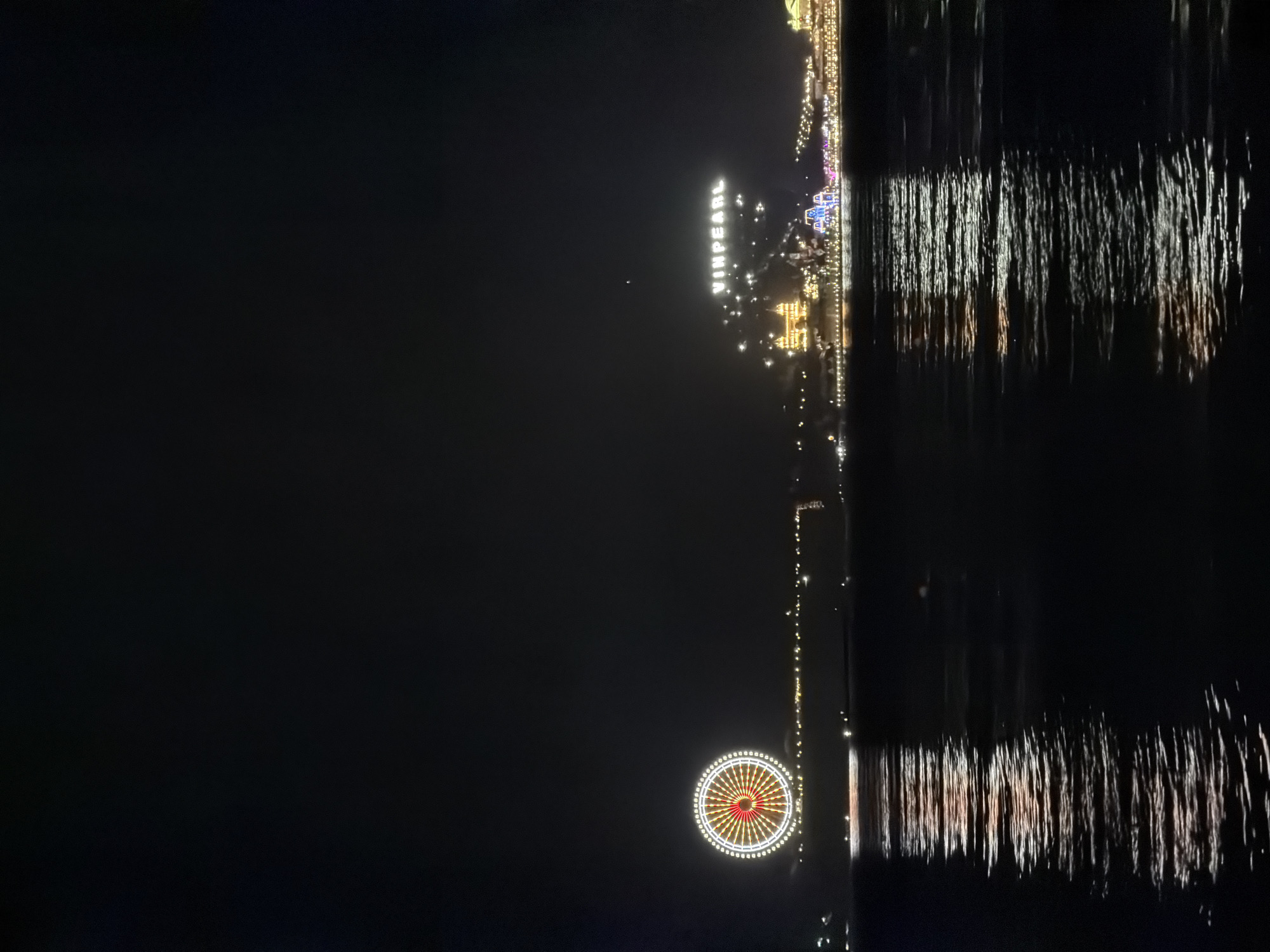 At night, a vertical view shows the illuminated shoreline of Nha Trang Ward, Vietnam, reflected in water. Prominently visible are a Ferris wheel with red and white lights, and the word "VINPEARL" spelled out in bright lights. Other structures along the shoreline are also illuminated with a variety of lights. The water surface exhibits bright streaks of light, reflecting the shore. The sky is dark and featureless.