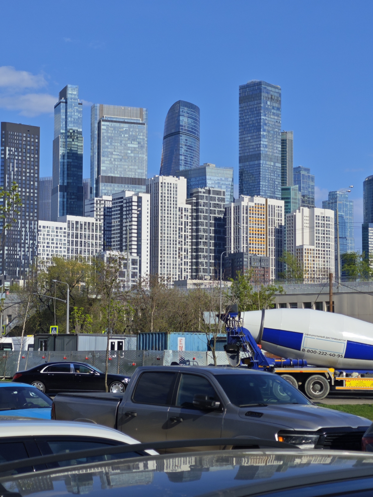 The image depicts a cityscape with a foreground dominated by parked cars, including a silver pickup truck. Behind the cars, there's a concrete mixer truck with a white and blue drum. A dark sedan is visible further back, near a fence with containers. In the background, a dense array of modern high-rise buildings and skyscrapers defines the Moscow International Business Center, also known as Moscow City. The sky is blue with scattered clouds. The location is Moscow, Russia. No people are clearly visible.