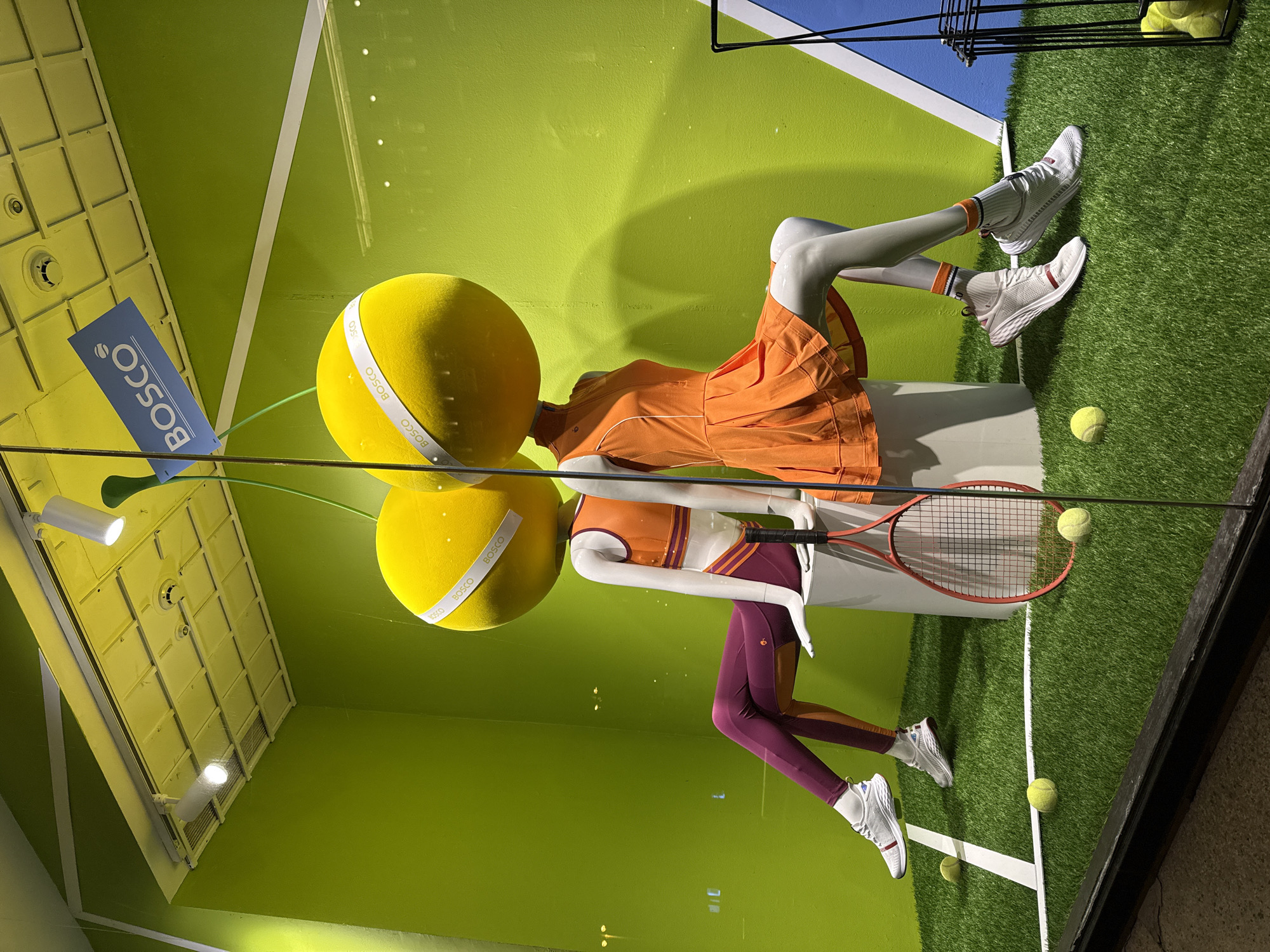 The image shows a store window display in Moscow, Russia, featuring tennis-themed mannequins. Two headless mannequins are dressed in tennis attire and arranged on a faux tennis court surface. The first mannequin wears an orange tennis dress with white shoes and socks. The second mannequin is dressed in a top and leggings, with tennis shoes. Each mannequin has a yellow ball with the word "BOSCO" printed on it as a head. The second mannequin holds a tennis racket. Several tennis balls are scattered on the artificial turf. The backdrop includes bright green walls with white lines mimicking a tennis court and a blue section at the top. A BOSCO logo is visible on a sign on the left side of the image. The ceiling structure is visible on the left side, along with several spotlights.