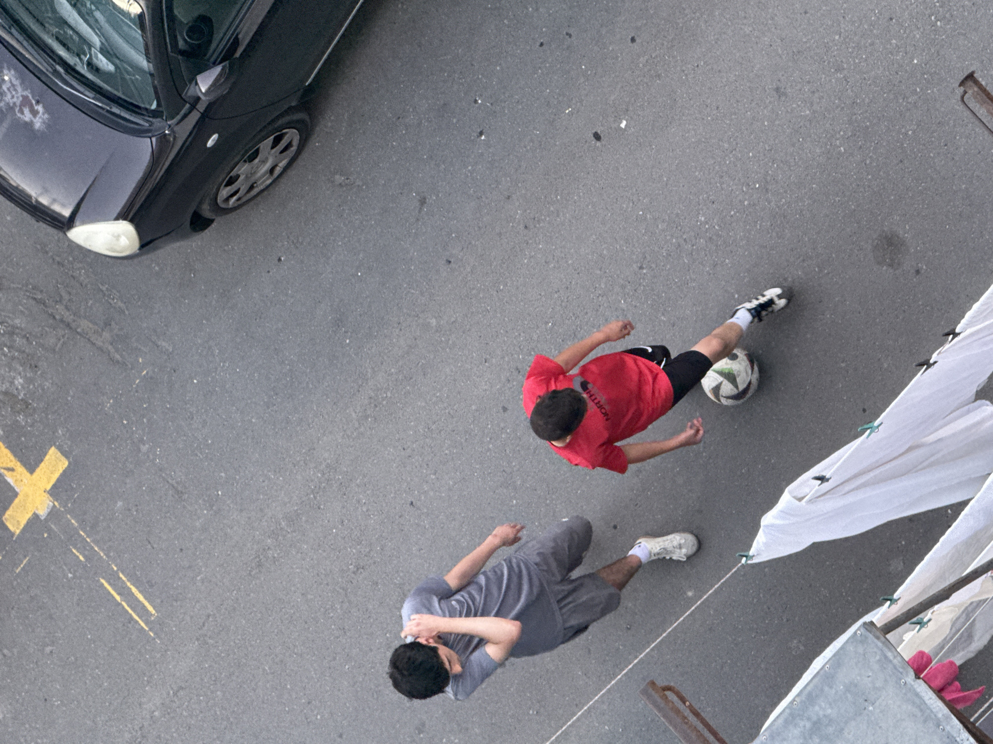 In Tbilisi, Georgia, two young males are captured in an overhead shot on an asphalt surface marked with yellow lines. The individual on the left, wearing a grey t-shirt and shorts, stands with his arm raised to his head. The other male, dressed in a red t-shirt and black shorts, is actively playing soccer with a ball at his feet. To the right of the frame, a black car is parked. In the foreground, a clothesline hangs with laundry, including white sheets and pink garments, secured with green clips. The scene appears to be a street or courtyard area.