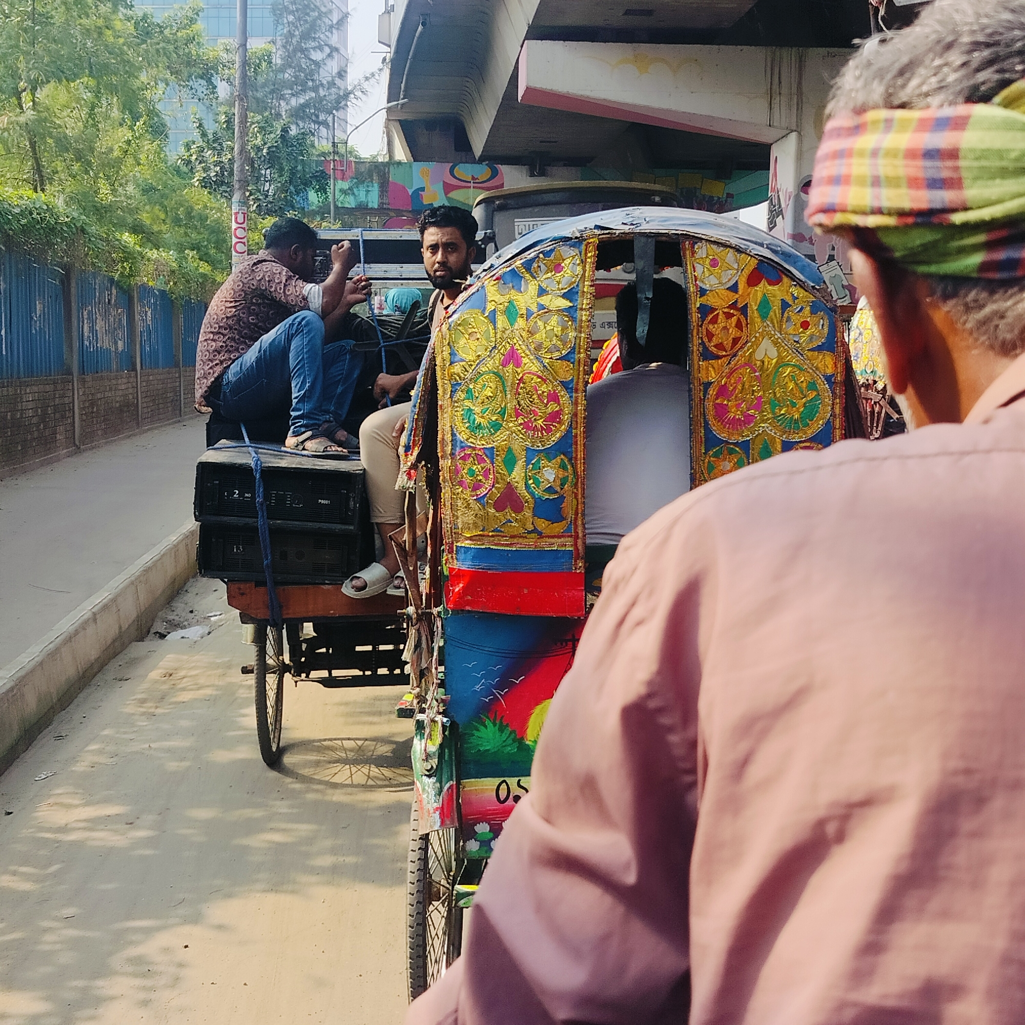 The image captures a scene on a street in Dhaka, Bangladesh, with a prominent cycle rickshaw elaborately decorated with colorful designs and patterns. On the back of the rickshaw, two men are seated atop what appears to be electronic equipment, possibly large speakers or amplifiers. One man is wearing a brown patterned shirt and jeans, while the other is wearing a light-colored top and trousers. A third man is visible in the rickshaw, possibly a passenger. The foreground features the back of a man wearing a pink shirt and a checkered headscarf. The street is lined with a concrete sidewalk and a fence on the left. Overhead, a portion of an elevated structure, such as a bridge or overpass, is visible. Buildings and trees can be seen in the background. The photograph captures a typical urban scene with the vibrant, decorated cycle rickshaws that are common in Dhaka.