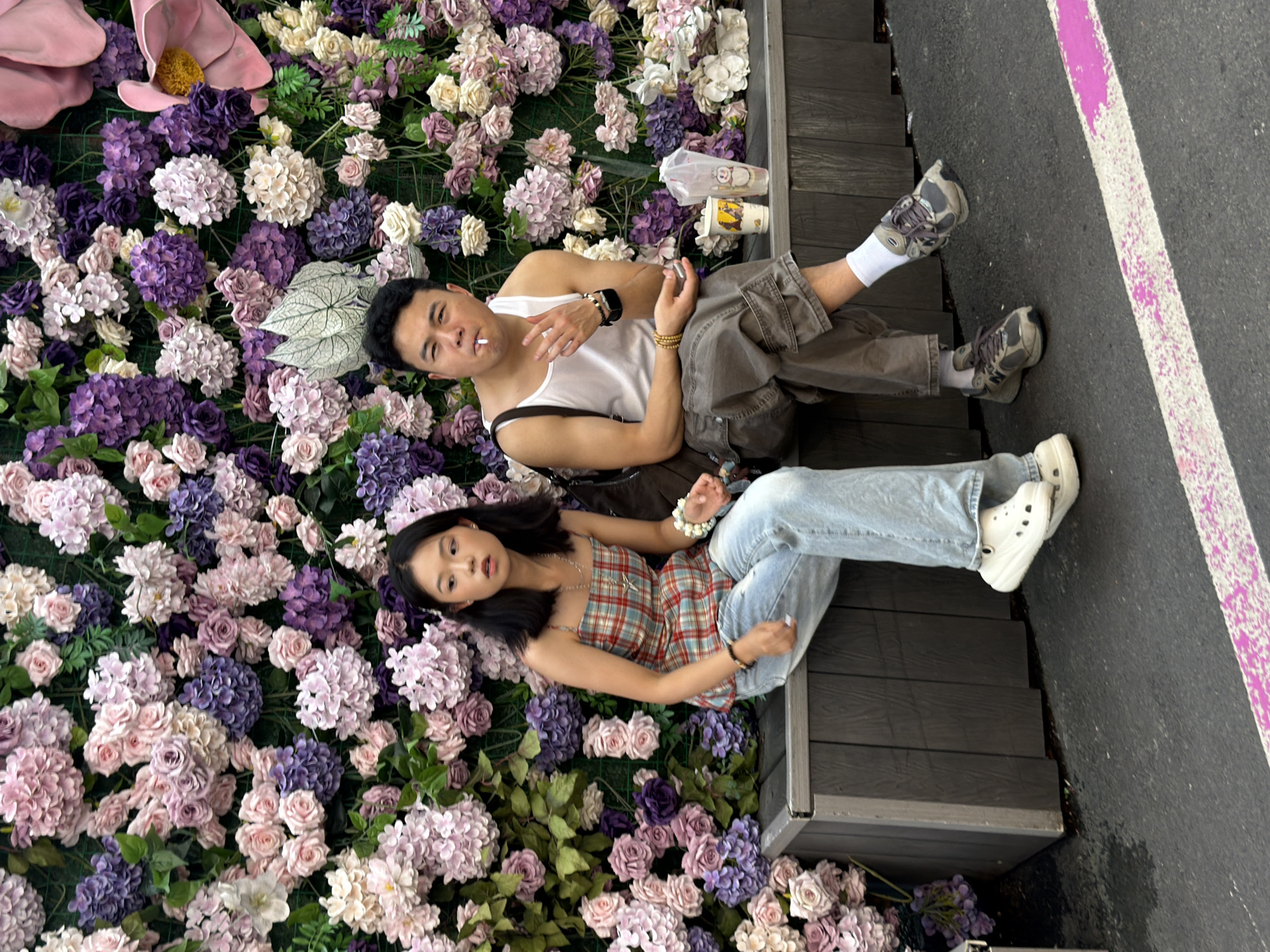 Two individuals are seated on a dark wooden bench against a dense backdrop of artificial flowers. The woman, positioned on the left, wears a plaid tank top, light-wash jeans, and white Crocs. She has dark hair and is looking towards the left of the frame. A beaded bracelet adorns her left wrist. The man, on the right, is wearing a white tank top, gray cargo pants, and athletic shoes with white socks. He has dark hair and is holding a cigarette in his mouth, with his right hand gesturing towards it. Next to him on the bench are two disposable cups. The scene appears to be outdoors, with asphalt and a pink and white stripe marking the ground in the foreground. The artificial flower wall features a variety of purple, pink, and white blossoms. The location is Yuzhong District, China.