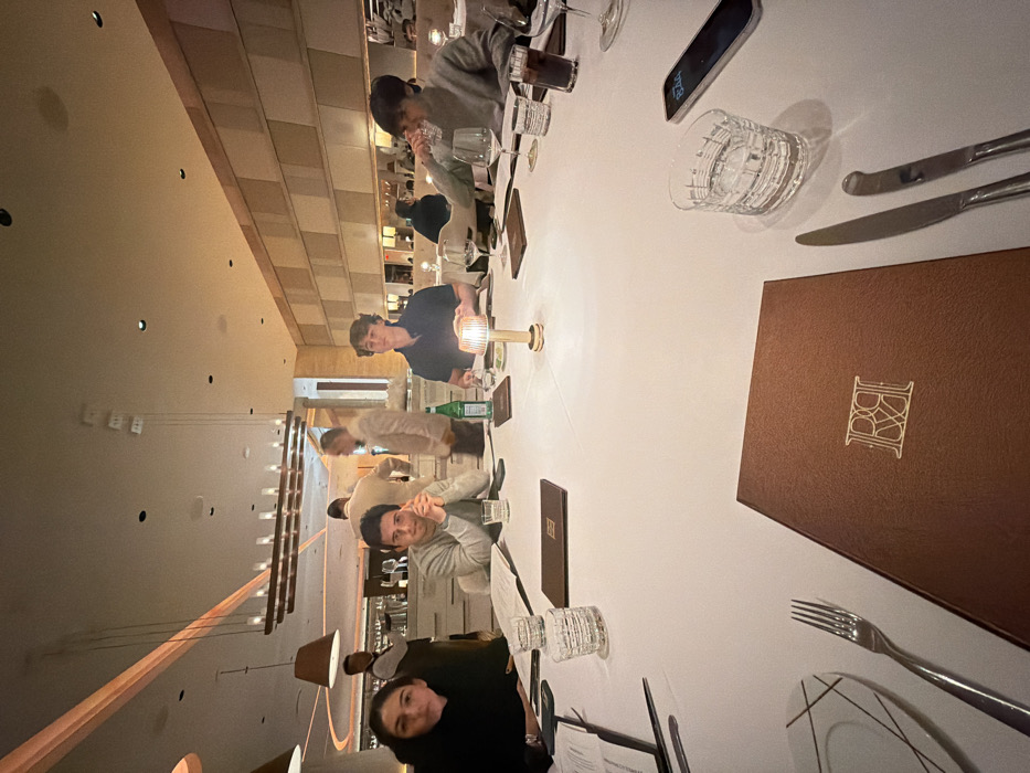A restaurant table is set with white linens, glasses, cutlery, and menus. People are seated around the table, appearing to be in a dining setting. A lit candle provides soft illumination. Modern lighting fixtures are visible in the background. The overall ambiance suggests a formal or upscale restaurant.