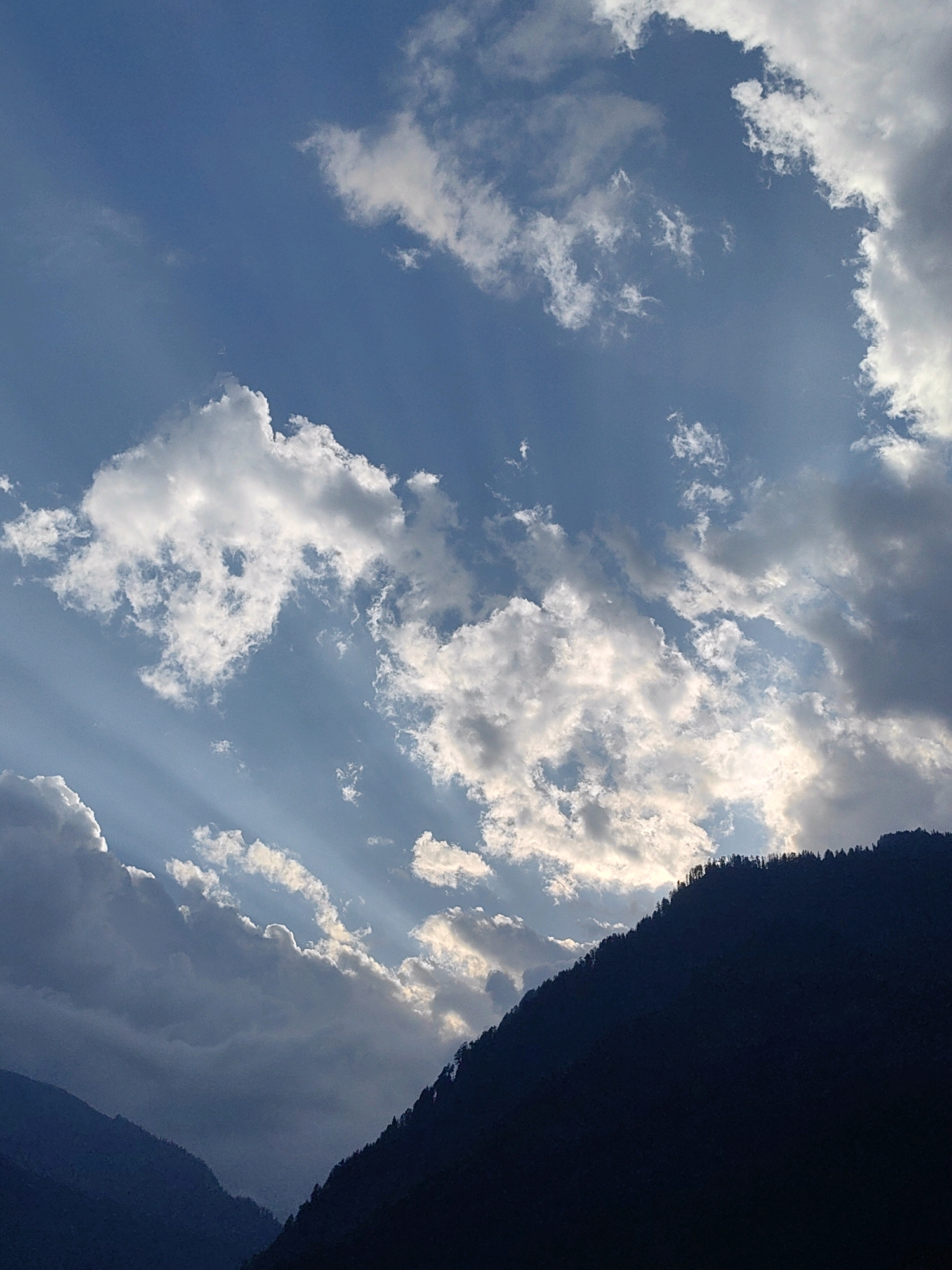 This is a photograph taken in Vashisht, India, capturing a dramatic sky over silhouetted mountains. The sky is a deep blue, filled with a mixture of bright white and darker grey clouds. Sunbeams are piercing through the clouds in several areas, creating shafts of light that illuminate the scene. The lower portion of the image is dominated by the dark, triangular shapes of mountains, their peaks outlined by the sky. The very top edge of the rightmost mountain is lined with the dark silhouettes of evergreen trees. The overall impression is one of a natural landscape bathed in dramatic lighting conditions.