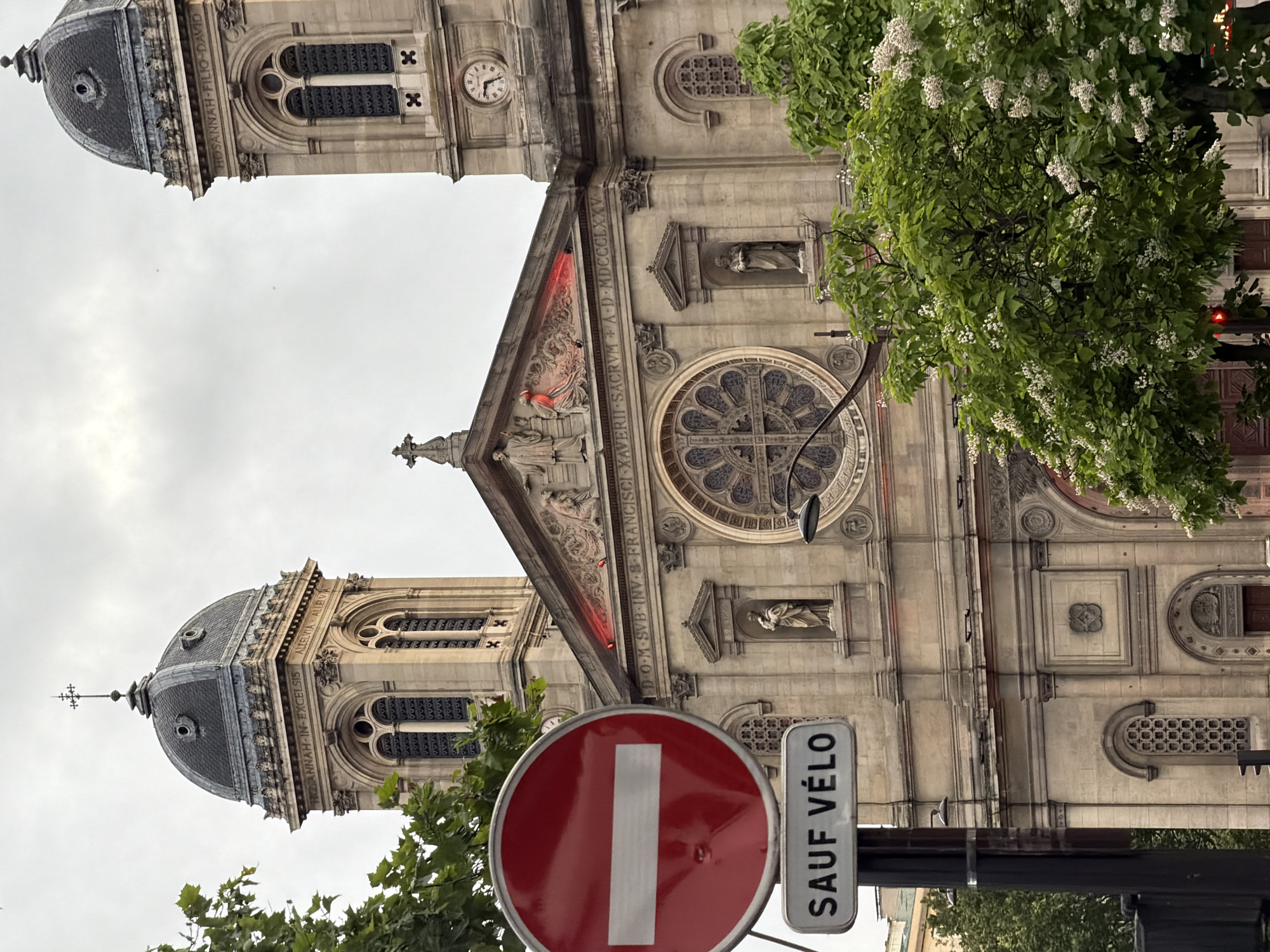 This image, captured in Paris, France, presents a rotated view of the grand facade of a historic church, likely Église Saint-François-Xavier. The impressive stone building features two prominent, symmetrical bell towers, each topped with a dark, domed roof and a spire. The architecture is ornate, displaying intricate carvings, arched windows, and statues positioned within niches.
The main facade includes a large, circular rose window with elaborate tracery. Above it, a central pediment is adorned with sculptural details. Latin inscriptions are visible across the building: "DOM SVB-INV-S-FRANCISCI-XAVERII SACRVM + A-D-MDCCCLXXV" is prominent above the rose window, identifying the church as dedicated to Saint Francis Xavier and built in 1875. On the tower to the right (as if the image were upright), the inscription "HOSANNAH-FILIO-DAVID" is visible, while the left tower displays "ROSANNAH-IN-EXCELSIS" and "ALLELVIA ALLELVA" (likely 'Alleluia'). A clock face showing a time just past ten is visible on the right tower.
Lush green trees, some bearing white blossoms, partially frame the building, suggesting springtime. In the foreground, a prominent red circular "no entry" sign with a white horizontal bar is visible. Beneath it, a white rectangular supplementary sign reads "SAUF VÉLO" (French for "except bicycles"), indicating that cyclists are permitted to enter despite the restriction for other vehicles. The sky is overcast, providing soft, diffused lighting to the scene. The image captures the architectural splendor of a Parisian church in an urban environment.