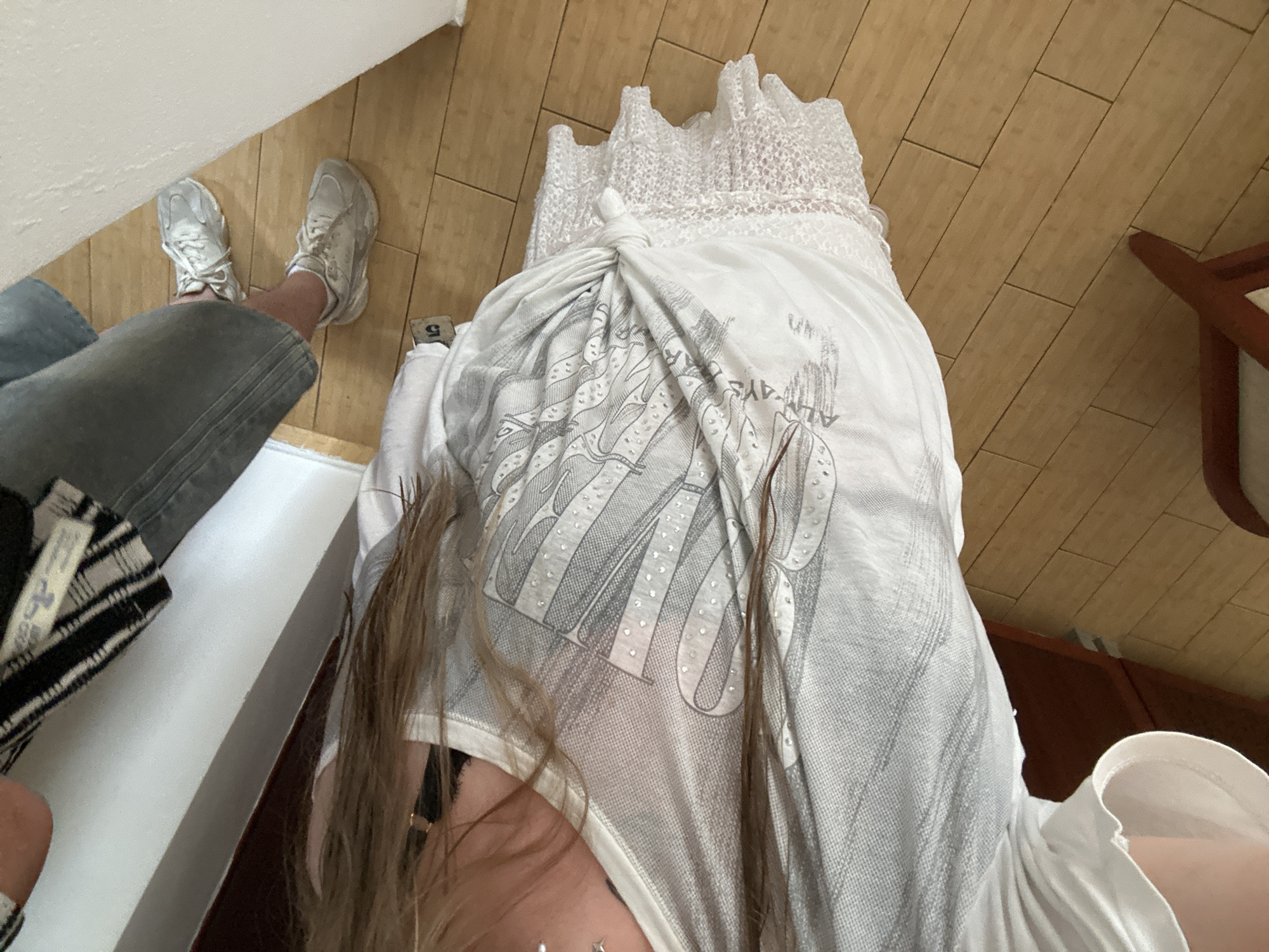 A high-angle shot captures a person in an indoor setting with a wooden floor, possibly in Bar, Montenegro (although this is not directly visible in the image). The person wears light grey jeans and grey sneakers. They are standing in front of a white garment that has been tied in a knot, partially obscuring a dress or similar item with a lace top underneath. The person is also wearing a white top with a grey pattern. A portion of their face and hair is visible. A wooden chair leg is visible to the right.