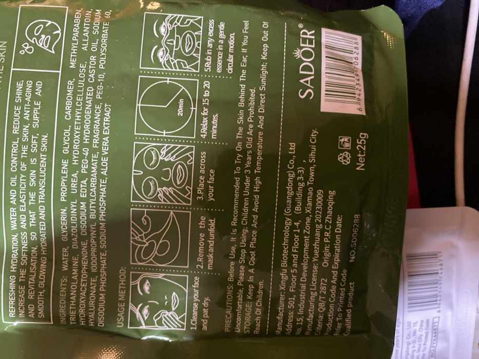 The image displays a close-up of a green, rectangular product package, likely for a face mask, oriented diagonally from the bottom left to the top right. The packaging is predominantly dark green with white text and line-art illustrations. The background is blurred, showing dark, indistinct shapes, possibly other items or fabric.
The front of the package is rich with information, divided into several sections:
1. **Product Benefits/Claims (Top Left):** A block of text describes the product's intended effects: "REFRESHING HYDRATION, WATER AND OIL CONTROL, REDUCE SHINE, INCREASE THE SOFTNESS AND ELASTICITY OF THE SKIN, ANTI-AGING AND REVITALISATION, SO THAT THE SKIN IS SOFT, SUPPLE AND SMOOTH, GLOWING HYDRATED AND TRANSLUCENT SKIN." An icon resembling a face with water droplets is next to this section.
2. **Ingredients List (Middle Left):** A detailed list of ingredients is provided: "WATER, GLYCERIN, PROPYLENE GLYCOL, CARBOMER, METHYLPARABEN, TRIETHANOLAMINE, DIAZOLIDINYL UREA, HYDROXYETHYLCELLULOSE, ALLANTOIN, HYDROXYACETOPHENONE, DISODIUM EDTA, PEG-40 HYDROGENATED CASTOR OIL, SODIUM HYALURONATE, IODOPROPYNYL BUTYLCARBAMATE, FRAGRANCE, PEG-10, POLYSORBATE 60, DISODIUM PHOSPHATE, SODIUM PHOSPHATE, ALOE VERA EXTRACT".
3. **Usage Method (Center Right):** Titled "USAGE METHOD:", this section presents five illustrated steps for applying the mask. Each step has a corresponding white line-art drawing inside a circle:
* "1.Cleanse your face and pat dry." (Illustration shows hands patting a face.)
* "2.Remove the mask and unfold." (Illustration shows hands unfolding a mask.)
* "3.Place across your face" (Illustration shows a mask applied to a face.)
* "4.Relax for 15 to 20 minutes." (Illustration shows a clock with "20min" and a relaxed face with closed eyes.)
* "5.Rub in any excess essence in a gentle circular motion." (Illustration shows hands rubbing the face.)
4. **Precautions and Storage (Bottom Middle):** This section outlines important warnings and storage instructions: "PRECAUTIONS: Before Use, It Is Recommended To Try On The Skin Behind The Ear, If You Feel Uncomfortable, Please Stop Using; Children Under 3 Years Old Are Prohibited. STORAGE: Keep In A Cool Place And Avoid High Temperature And Direct Sunlight; Keep Out Of Reach Of Children."
5. **Manufacturer and Product Information (Bottom Right):**
* The brand name "SADOER" is prominently displayed in large white letters with a crown-like logo above it.
* A barcode is visible to the right of the brand name, with the number "6942349706288".
* "Net:25g" is indicated, accompanied by two recycling symbols.
* Detailed manufacturer information: "Manufacturer: Xingfu Biotechnology (Guangdong) Co., Ltd", "Address: 501, Floor 5 and Floor 1-4, (Building 3-3) No. 15, Industrial Development Zone, Xiamao Town, Sihui City."
* Further details include: "Manufacturing License: Yuezhuang 20230009", "Criteria: QB/T 2872", "Origin: P.R.C Zhaoqing", "Production Code And Expiration Date: Refer To Printed Code", "Qualified product", and "NO.SD06288" (a product number).
The overall impression is a commercially packaged cosmetic product with comprehensive instructions and information printed directly on the pouch.