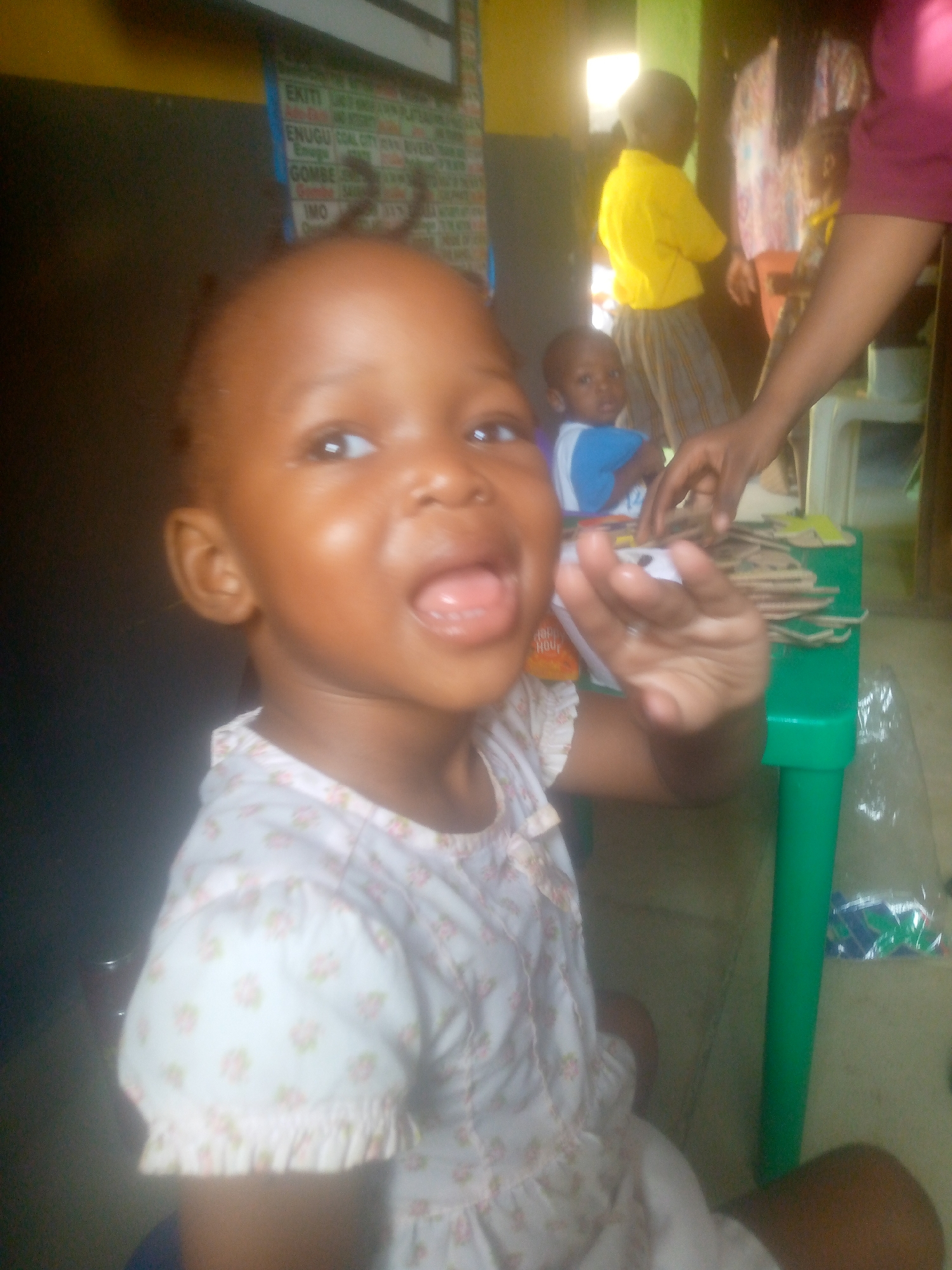 A young child with dark skin and short, curly hair is the main subject of the photo. The child is wearing a light-colored dress patterned with pink flowers. The child's mouth is open in a smile or shout, and one hand is raised slightly in the air.
In the background, several other children are visible. One child wears a yellow shirt and grey pants, and another wears a blue and white shirt. An adult hand reaches toward a pile of cardboard shapes on a green table. A green table leg is visible. Other details of the room include a dark wall to the left, a poster or sign with text on the wall, and additional furniture or objects in the background.
Given the text on the poster (including "ENUGU" and "IMO") and the image context, the location is likely a school, daycare, or similar setting in Ikorodu, Nigeria.