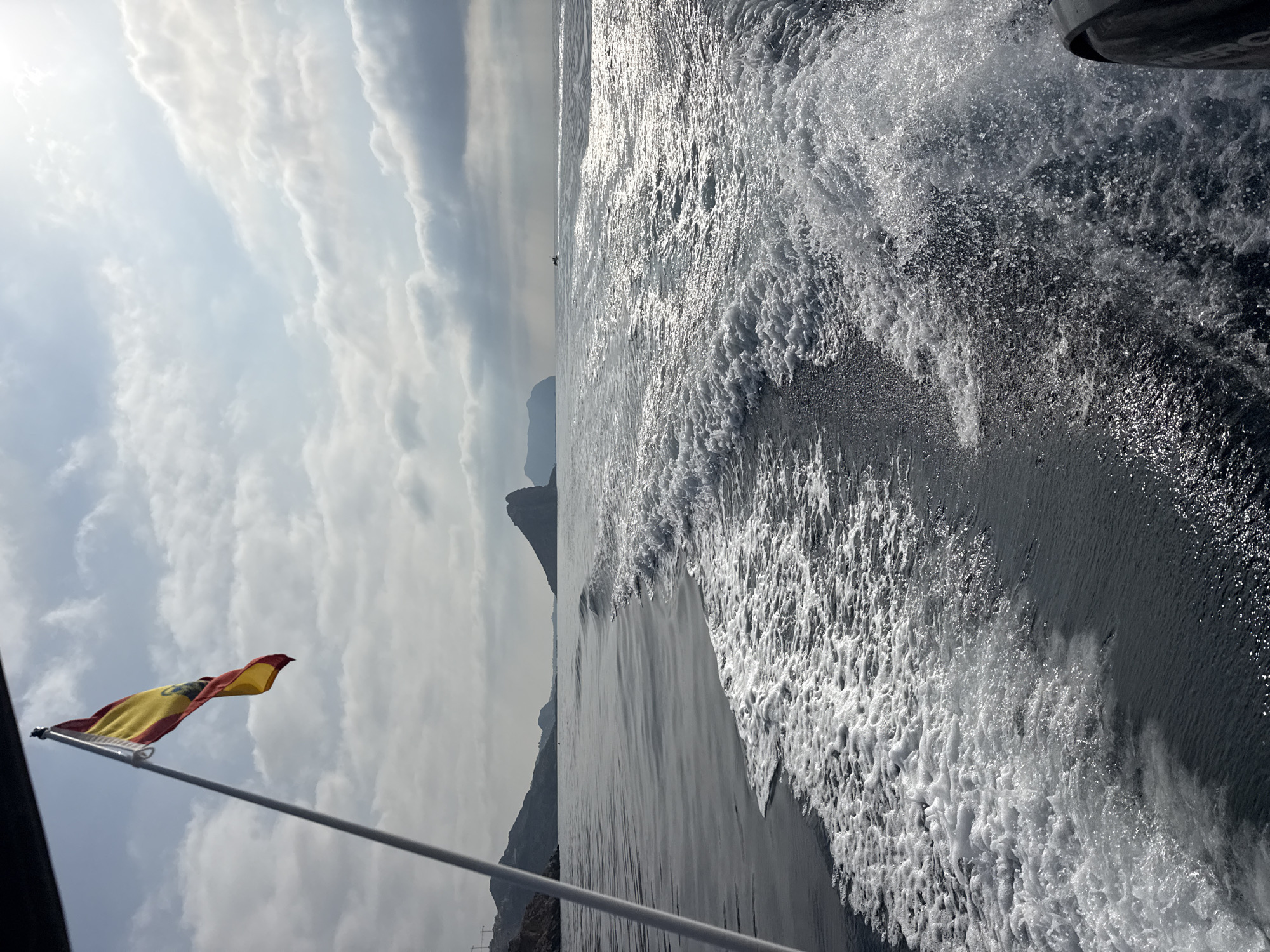 A Spanish flag flutters from a pole on a boat moving through the water, creating a prominent wake. The boat is traveling away from the shore, which is lined with rocky hills and mountains. The sky is partly cloudy with bright sunlight filtering through. In the distance, there are a few other boats visible on the sea. The scene captures a day out on the water, likely near Águilas, Spain, given the prominent Spanish flag and the coastal landscape.
