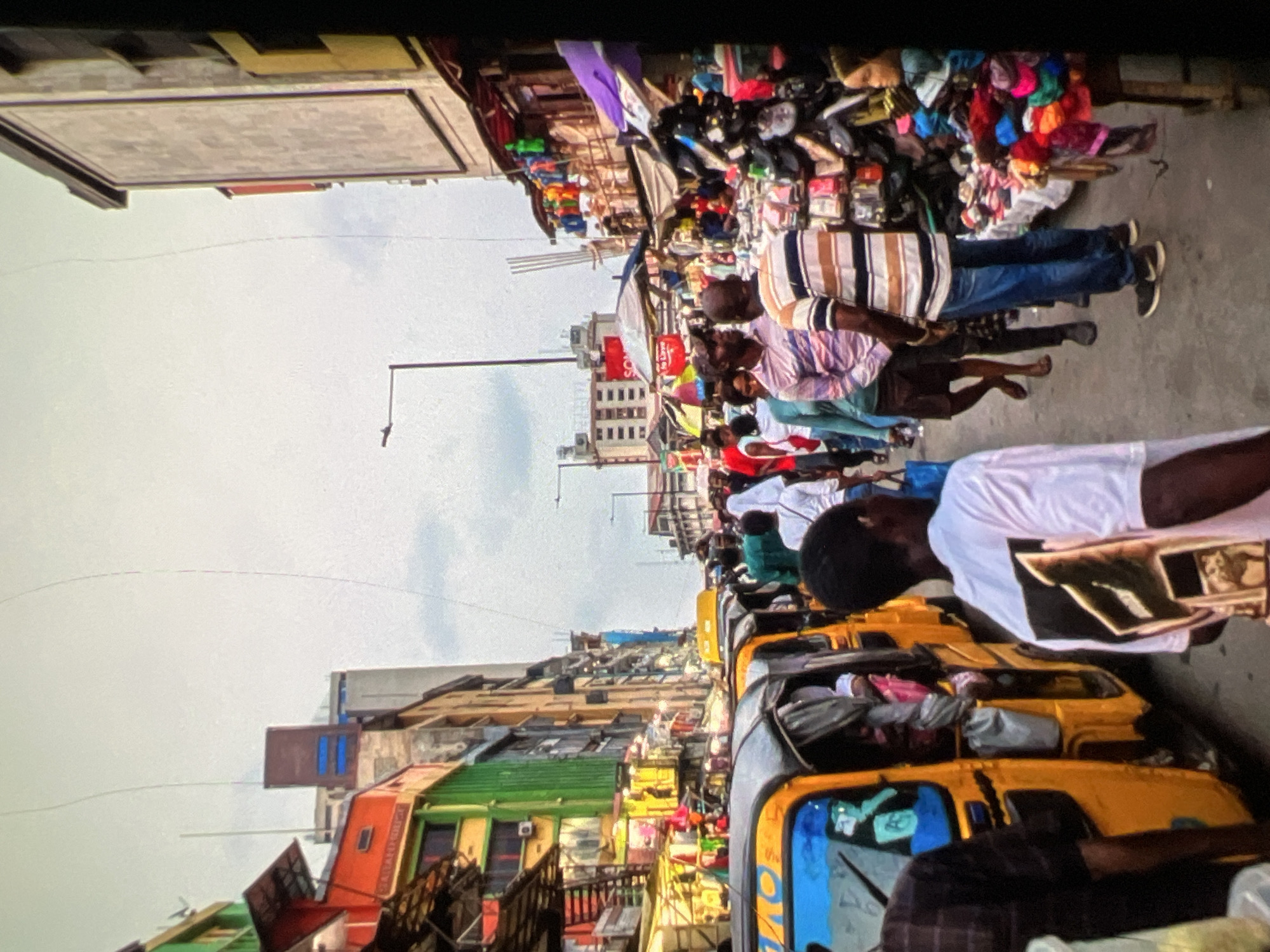This image captures a bustling street scene in Lagos, Nigeria. A narrow street is packed with people and vehicles, suggesting a busy marketplace or thoroughfare. On the left side, a row of colorful buildings, predominantly green and orange, line the street. These structures appear to be multi-story shops or residences, with visible windows and balconies.
In the foreground and extending down the street are several yellow auto-rickshaws, commonly known as 'keke napep' in Nigeria. These are filled with passengers and luggage, indicating active transportation. Pedestrians are walking in both directions, navigating through the crowded space. They are diverse in age and gender, wearing casual clothing typical of a tropical climate.
To the right of the street, market stalls are set up. These stalls display a variety of goods, including textiles, clothing accessories, and what appear to be toiletries or beauty products. A mannequin head wearing a headscarf is visible among the merchandise. The stalls have awnings and umbrellas for shade.
In the background, more buildings of varying heights and architectural styles can be seen, some appearing modern with glass facades, while others are older and more traditional. Streetlights are present, and overhead wires crisscross the sky, a common sight in urban Nigerian environments. The sky is overcast, suggesting a cloudy day. The overall impression is one of vibrant, energetic urban life, characteristic of a busy commercial area in Lagos.
