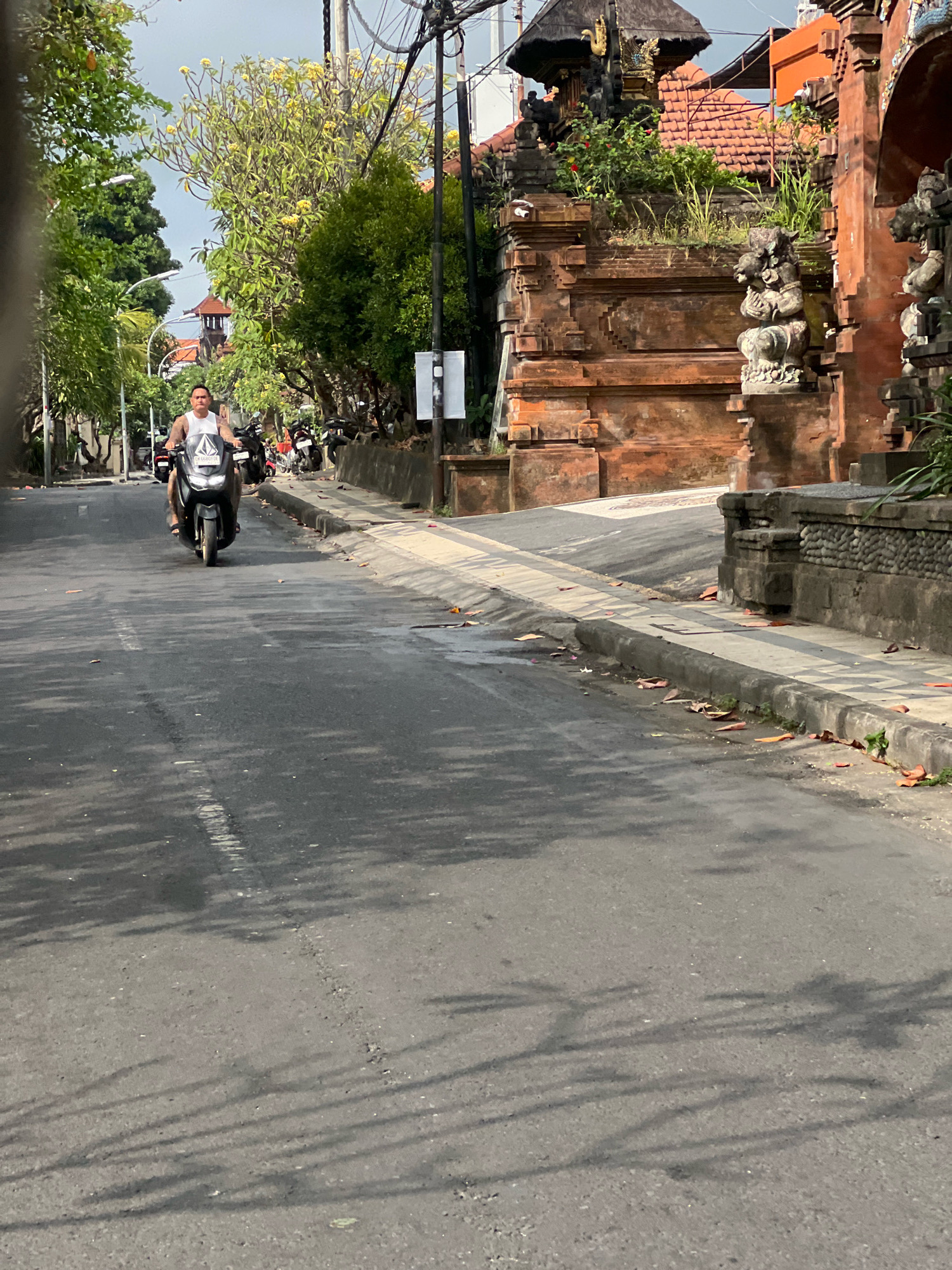A man rides a dark-colored scooter down a paved road in Kuta, Indonesia. The man is wearing a white tank top with a graphic logo and dark shorts. He has tattoos on his arms. The scooter has a dark windshield. On the right side of the road is a traditional Balinese building with intricate stone carvings and a tiled roof. The building is made of orange brick and stone. Several other scooters are parked on the side of the road near the building. Trees with green leaves and some yellow flowers line the left side of the road. The sky is overcast. The road has a white painted line down the center and some fallen leaves on the asphalt. Shadows of trees are cast across the road.