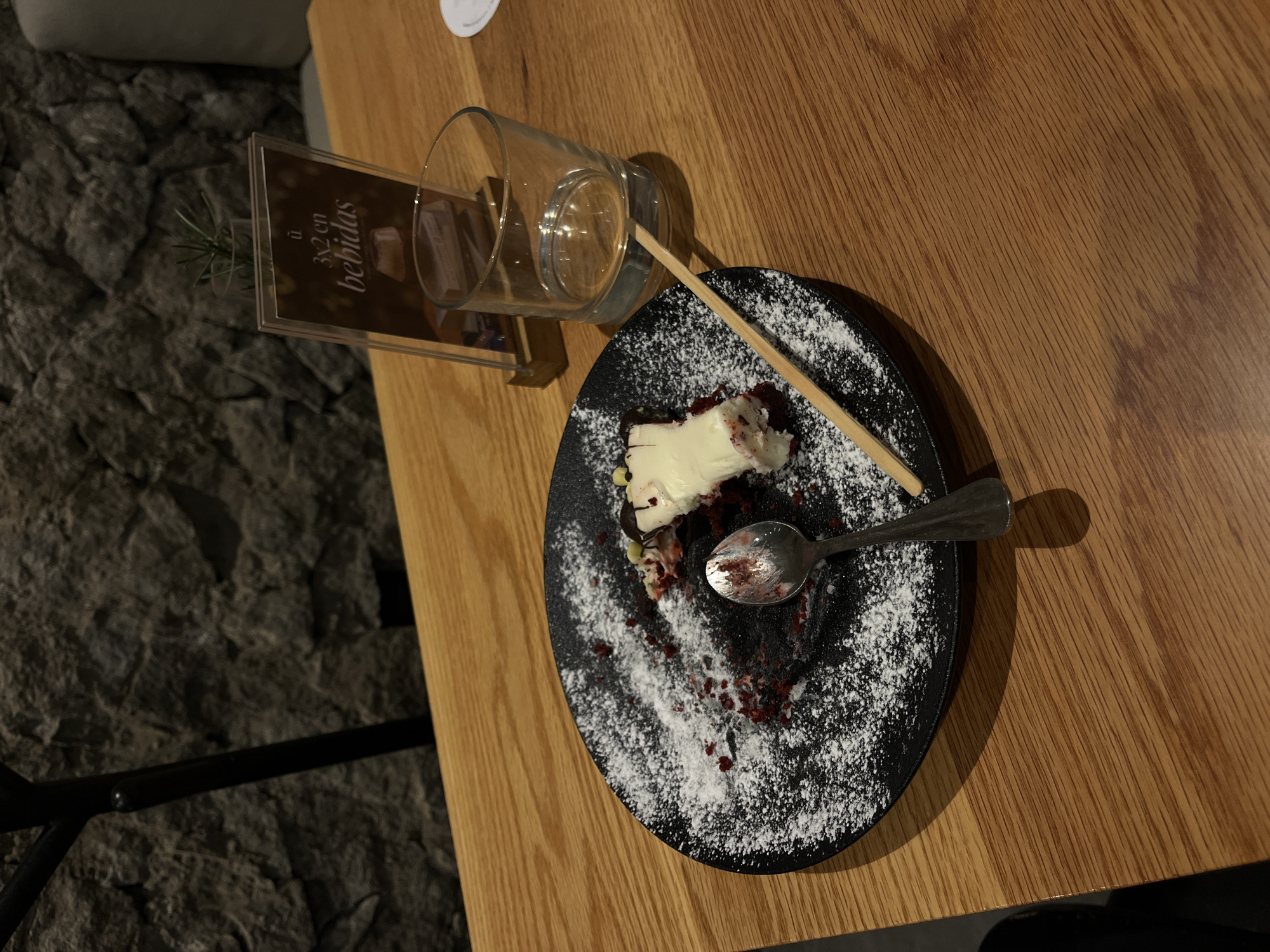 The image shows a partially eaten dessert on a black plate dusted with powdered sugar. A spoon rests on the plate, its bowl stained with remnants of the dessert. A wooden skewer also lies across the plate. Next to the plate is a drinking glass and a small promotional display stand for "3x2 en bebidas," indicating a drink special. The setting is a wooden table. The location is San Bernardino Tlaxcalancingo, Mexico. No people are visible.