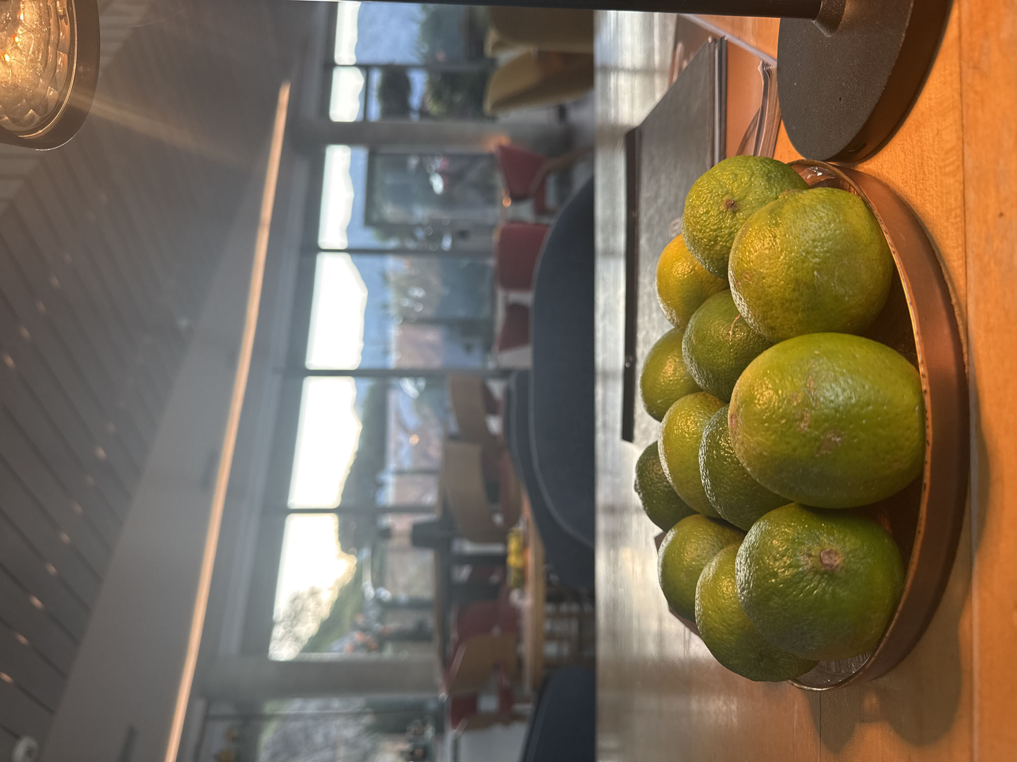 The image depicts a bowl of limes on a wooden surface, likely a table or countertop, with a blurred background featuring a well-lit interior and a view through large windows. The location is plausibly Đurići, Montenegro, suggested by the landscape visible through the windows, possibly showing a mountainous region and bright sky. There are no people clearly visible, but there are chairs, a table, and a yellow bench in the background that has been out of focus, suggesting the interior could be a cafe or a restaurant. The lemons are clustered together in a round metal bowl. The lighting is bright, with an overhead lamp visible in the corner. The scene appears to be indoors during daylight.