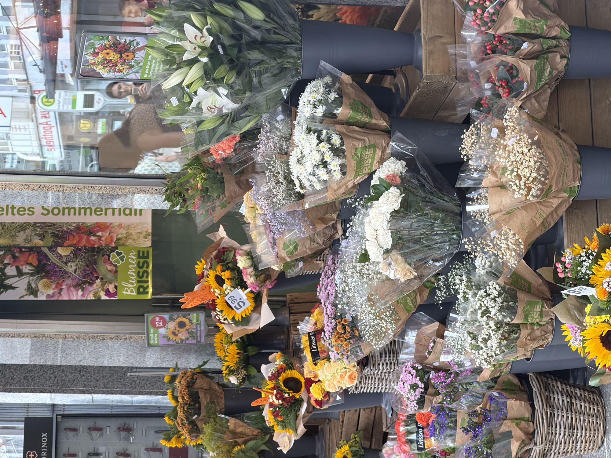 This image depicts a vibrant outdoor flower stand located in front of a shop with a large glass window, likely in the city of Witten, Germany. The shop's facade features a grey, patterned stone exterior. A green banner with the text "BLUMEN RISSE" and a four-leaf clover logo is prominently displayed. The flower stand itself is overflowing with a diverse assortment of flowers arranged in bouquets and bunches, many of which are wrapped in brown paper and clear plastic.
Sunflowers are a dominant feature, with several large bouquets of them visible, some priced at €12.99 and €5.99 respectively. Other flowers include vibrant yellow chrysanthemums, delicate white baby's breath, pink and white roses, and bouquets of small purple and pink blossoms. Tall, elegant white lilies in a large grey vase stand out in the background. A wicker basket filled with smaller floral arrangements is also present.
Reflected in the shop's glass window, which displays various advertisements and a bus stop sign, are blurred figures of people, suggesting pedestrian traffic. One woman is clearly visible inside the shop, looking at her phone. The overall scene conveys a lively and colorful market atmosphere.