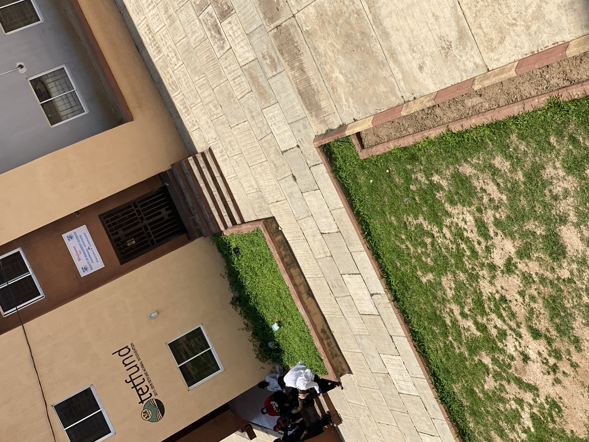 This image shows a high-angle view of a building facade and an outdoor area, likely in Jos, Nigeria. The building is painted in neutral tones of beige and brown. Several windows with dark panes are visible on the facade. A sign on the building reads "Welcome to FACULTY LIBRARY FACULTY OF MANAGEMENT SCIENCES JOURNAL OFFICE". Below this, a larger logo and text indicate "tetfund 2017-2019 TETFUND NORMAL INTERVENTION".
In front of the building, a group of people are gathered under an overhang. They are dressed casually, with some wearing white. The activity appears to be a meeting or a casual social gathering.
The outdoor area features a paved walkway made of rectangular stones. Adjacent to the walkway is a strip of green grass, bordered by a raised planter with reddish-brown and yellow edging. A larger grassy area, with patches of dry earth, extends beyond the planter. The scene suggests a sunny day, indicated by the shadows cast by the building and the people. The overall setting is that of an academic or institutional campus.