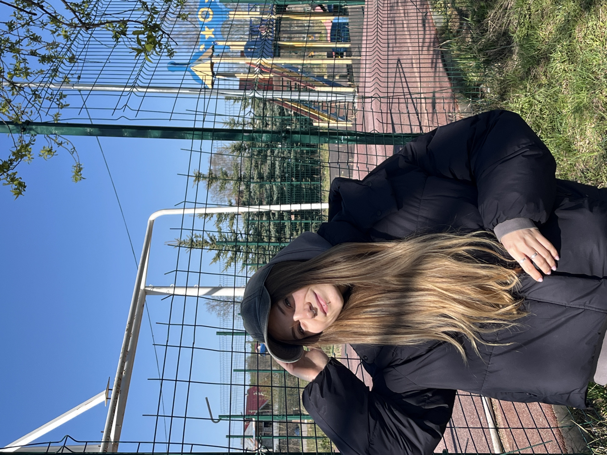 The image depicts a woman standing in front of a green wire fence. In the background is a playground with a slide and other equipment. The location is likely Фоки, Russia, based on the user prompt. The woman is wearing a black hooded jacket and a baseball cap. Her hair is long and blonde. She is holding the brim of her cap. The day appears sunny with a clear blue sky. There is greenery visible beyond the fence.