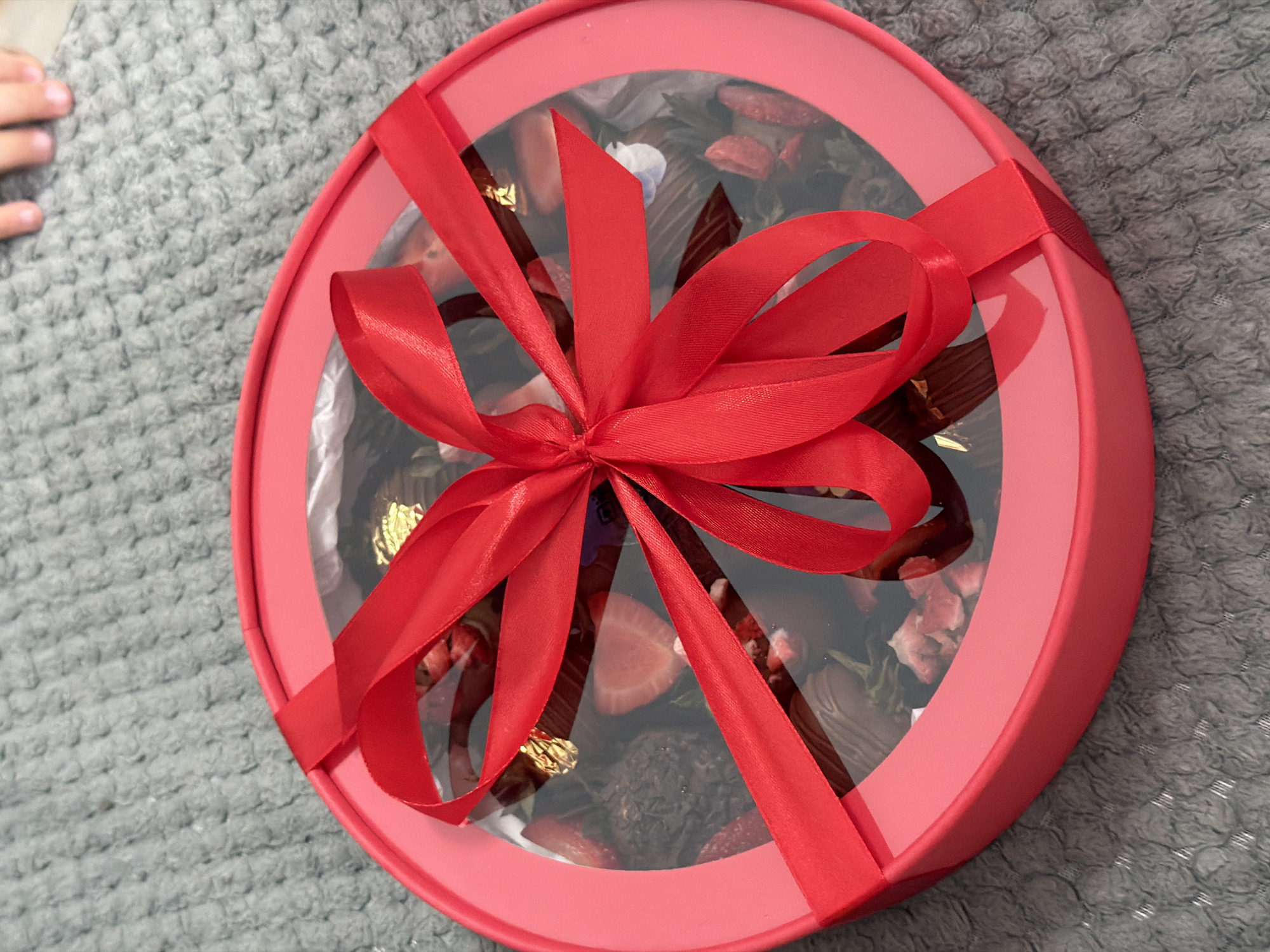 A red heart-shaped gift box containing chocolates and strawberries is placed on a grey, textured surface. The box has a clear lid, allowing the contents to be seen. A bright red ribbon is tied in a bow around the box. The background shows a hint of a child's fingers at the top right, suggesting a domestic setting. The image does not contain any identifiable landmarks or features that would allow for its location to be determined within the city of Chaikovsky, Russia.