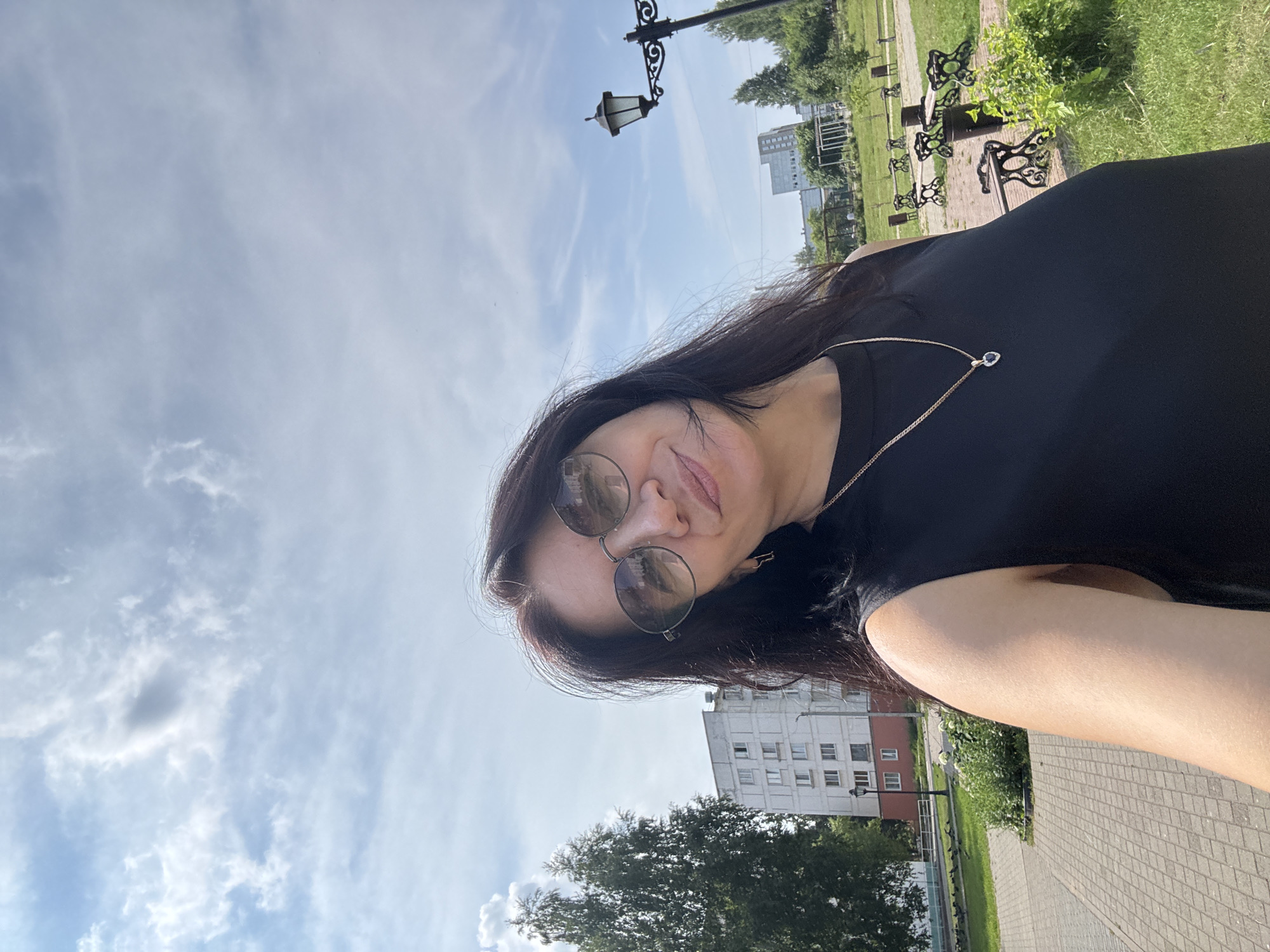 A woman with dark hair and sunglasses takes a selfie outdoors in Chaikovsky, Russia. She is wearing a black sleeveless top and a gold necklace with a heart-shaped pendant. Behind her, a paved walkway leads to a grassy area with park benches, a lamppost, trees, and apartment buildings in the background. The sky is blue with scattered clouds.