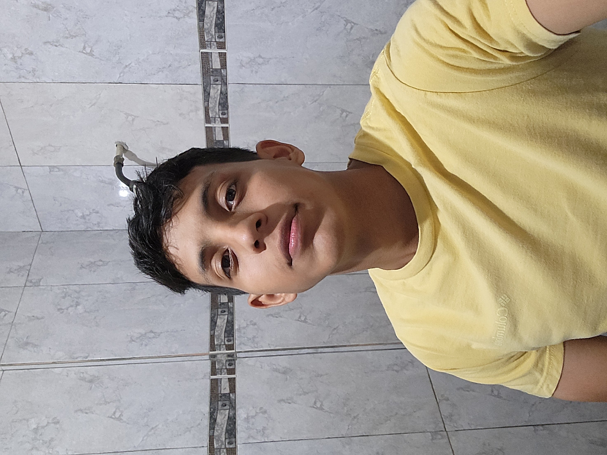 A young male is depicted indoors, possibly in a bathroom, as suggested by the tile-lined wall in the background. He is wearing a yellow t-shirt with "Columbia" printed vertically in white on the lower right side. A black pipe is seen protruding from the wall behind the top of his head. The image does not provide specific information to pinpoint a location in Tinaquillo, Venezuela. The focus is on the individual's portrait within the enclosed space.