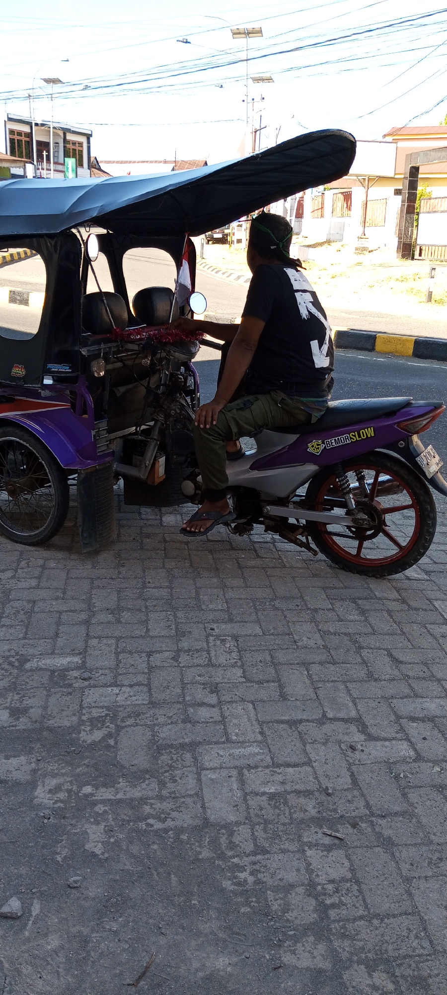 A man is seated on a purple and grey three-wheeled motorcycle, facing away from the camera, in Pakkanna, Indonesia. He is wearing a black t-shirt with white lettering and dark green cargo pants, with a green bandana tied around his head. The motorcycle has a black canopy extending over the seating area. The man's left hand rests on the handlebars, and his legs are positioned as if preparing to drive. The motorcycle is parked on a paved walkway made of grey interlocking bricks, adjacent to a road with yellow and black striped speed bumps. In the background, buildings with white walls and tiled roofs are visible, along with utility poles and overhead wires. The sky is bright and clear.