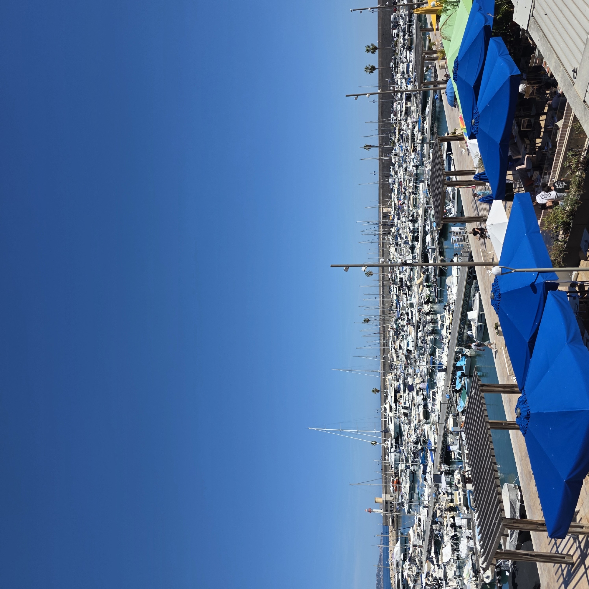 This is an image taken in the city of Menton, France, showing a marina filled with numerous boats and yachts under a clear blue sky. In the foreground, several open-air cafes are visible, with blue and green umbrellas providing shade over tables and chairs. Some people are seated at the tables, while others are walking or standing nearby. In the middle ground, wooden pergolas offer further shade, and a pathway runs along the marina's edge where people are seen strolling. Beyond the cafes and pathway, the marina is densely packed with a variety of boats, ranging from smaller vessels to larger yachts, all docked in the tranquil blue water. In the background, a concrete pier with a lighthouse at its end stretches into the sea, and beyond that, the coastline of Menton is faintly visible, with buildings and a few palm trees dotting the landscape. The overall scene depicts a sunny day at a bustling French Riviera harbor.
