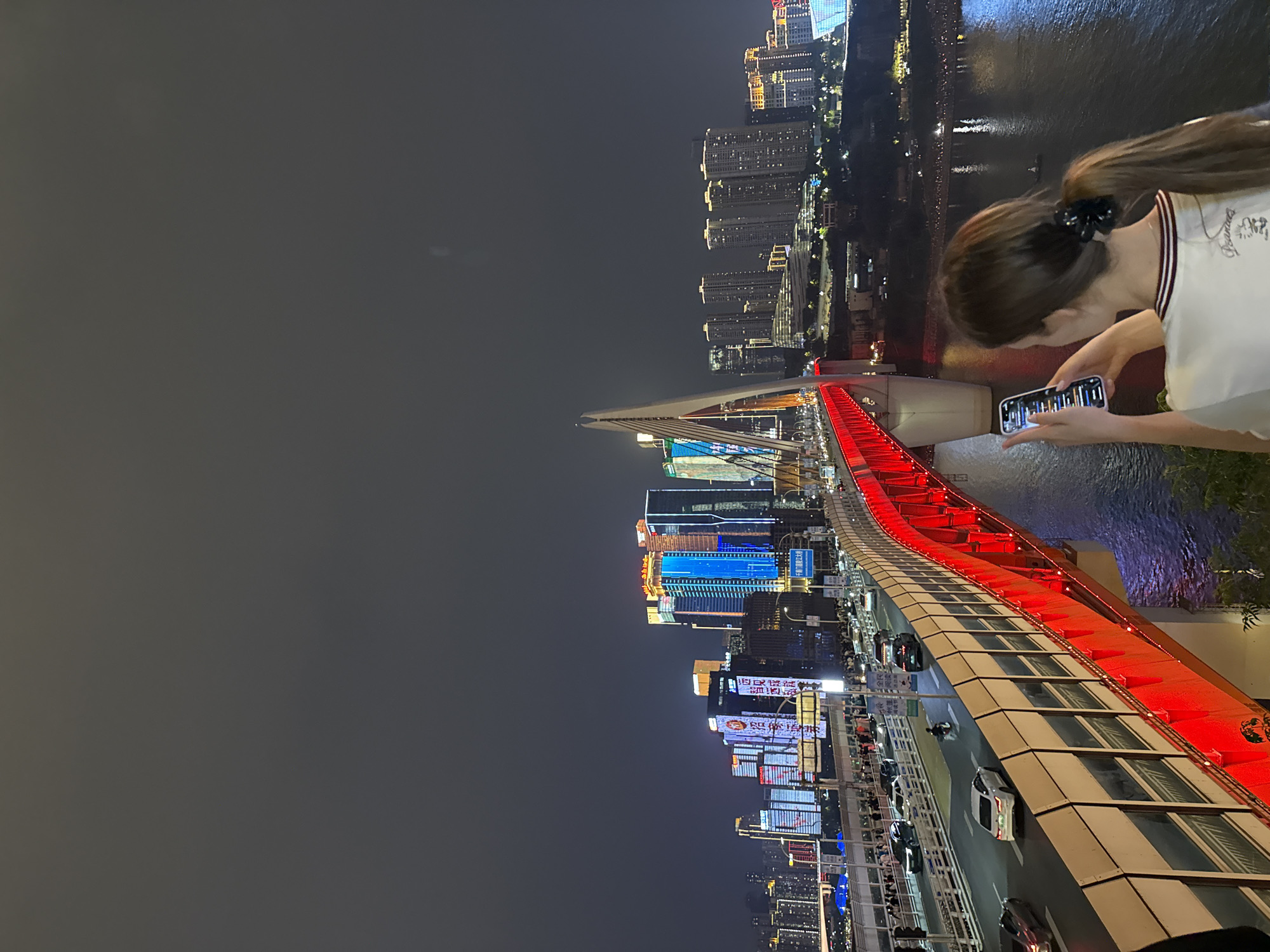 At night, a woman stands on the Hongyadong Scenic Area in Yuzhong District, China, holding and looking at her smartphone, with a red-lit bridge visible to the right, and city buildings in the background.