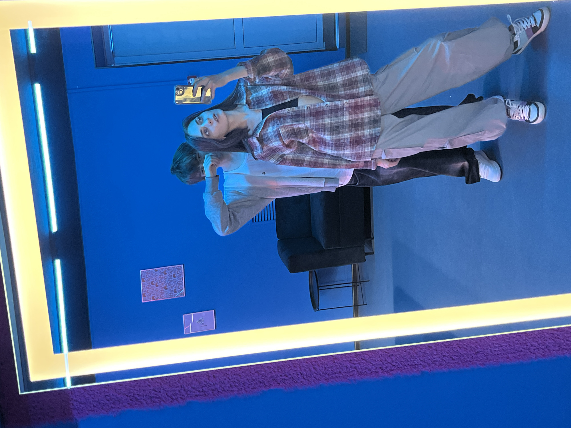 A mirror reflects two young individuals in a room setting. The foreground of the mirror reveals a frame with illuminated edges.
The individual on the right is holding a mobile phone, seemingly taking a photograph or video of themselves and another person. They are clad in a flannel shirt, black top, and light-colored pants, with white sneakers.
The other individual is wearing a light-colored shirt and dark pants, with white shoes.
Behind the figures, in the reflected background, there's a couch, a chair and two picture or photos on the wall.
The scene is set against a blue wall with a blue floor and a purple, furry object at the bottom of the image. The location is Krasnoyarsk, Russia.