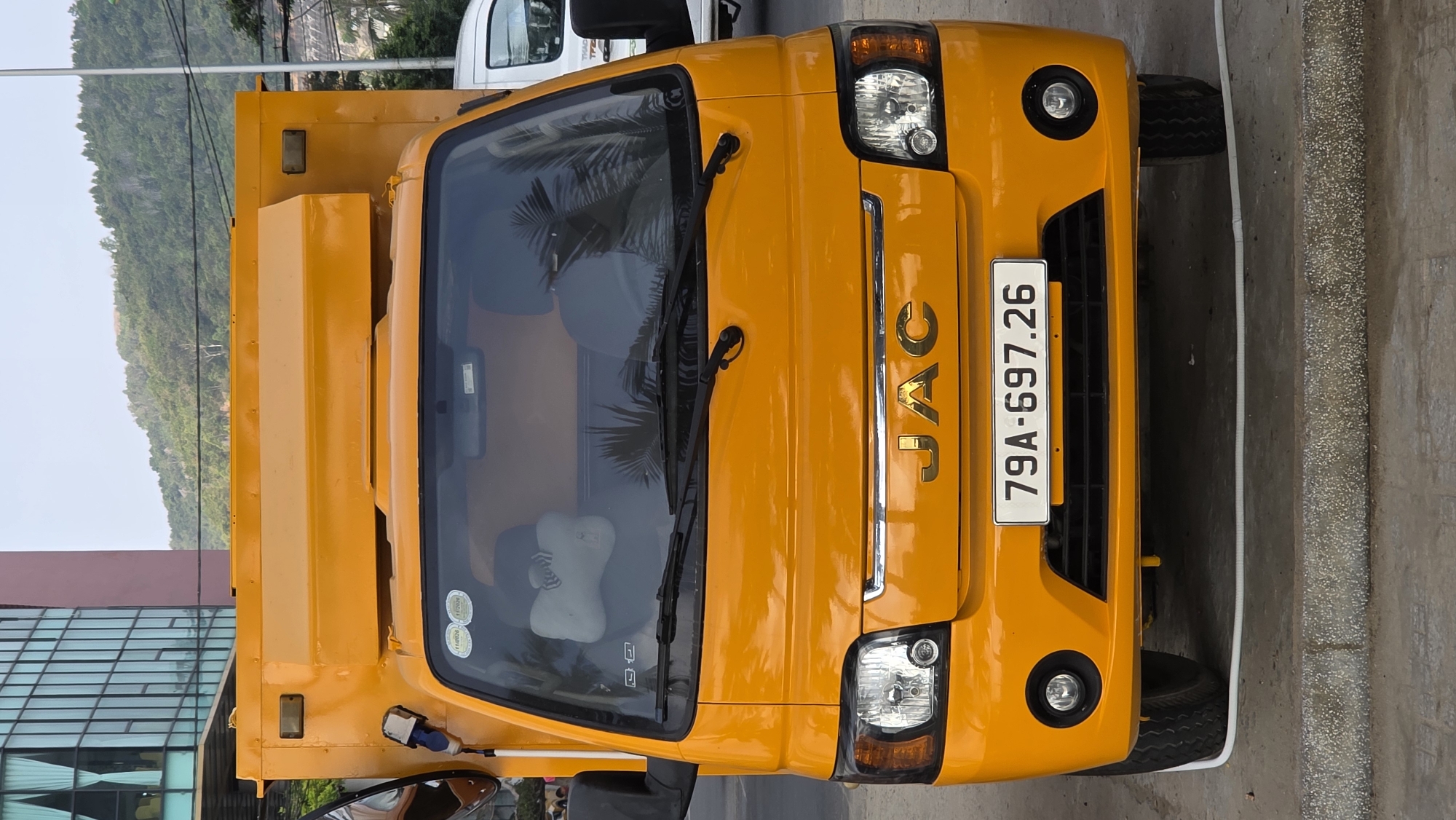 A yellow JAC truck is parked in Bac Nha Trang Ward, Vietnam. The license plate number is 79A-697.26. The truck faces the camera. A building with glass windows and a tree-covered hill are in the background. Another vehicle is partially visible behind the truck.