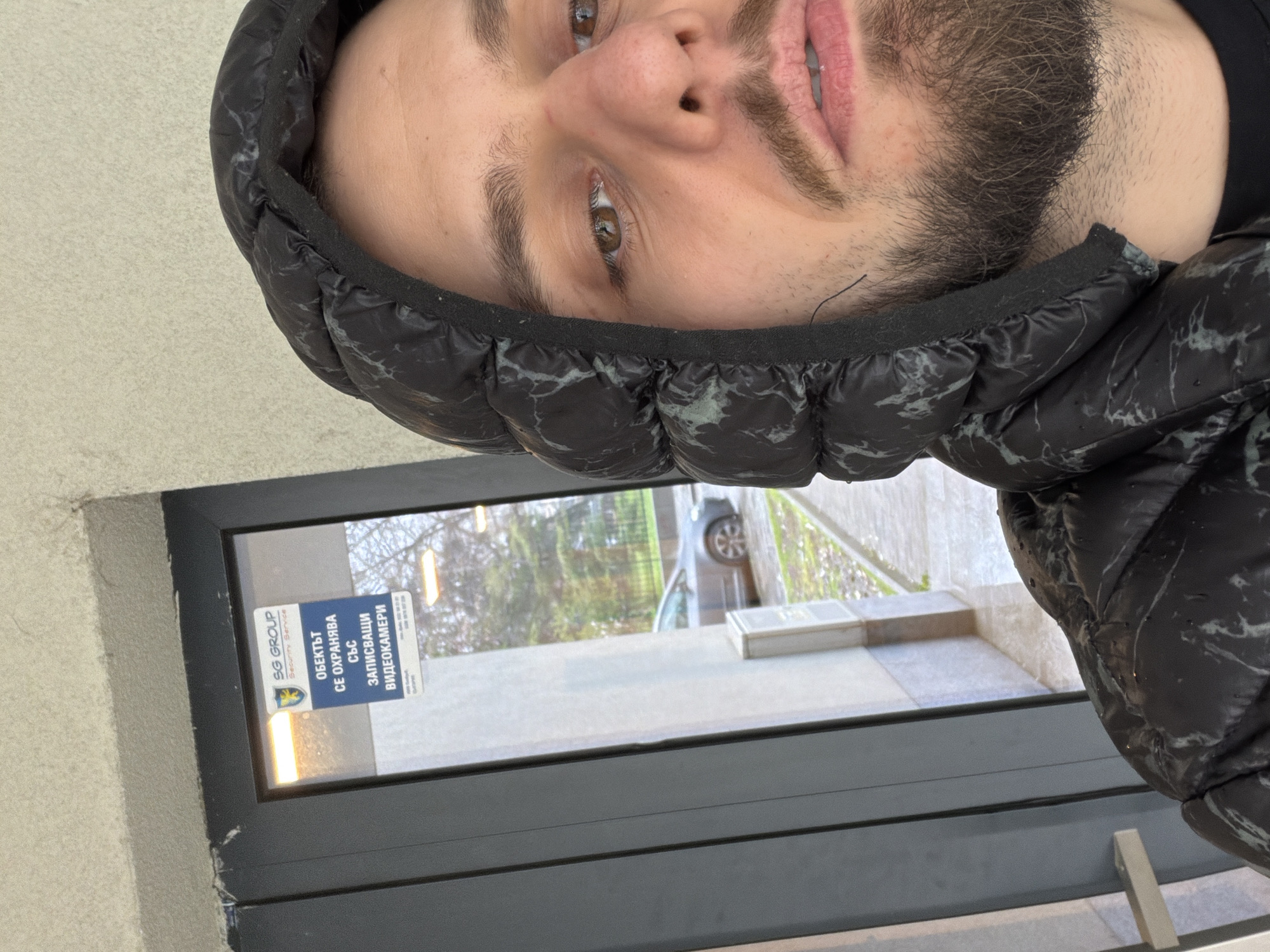 A man with a beard and a black puffy jacket with the hood up is positioned at an angle, likely taking a selfie. Behind him is a window with a sticker in Bulgarian that includes the words "SG GROUP" and "ВИДЕОКАМЕРИ," indicating video surveillance. The view through the window shows an outdoor scene with trees, a car, and a paved area. Based on the style of the sign and general look of the urban environment, this is potentially located in Plovdiv, Bulgaria.