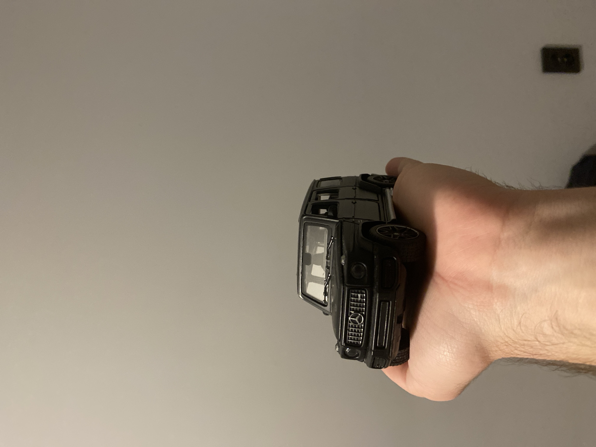 A person's hand holds a black toy Mercedes-Benz G-Class SUV model. The toy car is positioned in the center of the image, resting on the person's palm. The background is a plain, light gray wall with a dark electrical outlet visible on the lower right side. There is no discernible indication of the city of Łódź, Poland, or any specific urban environment.