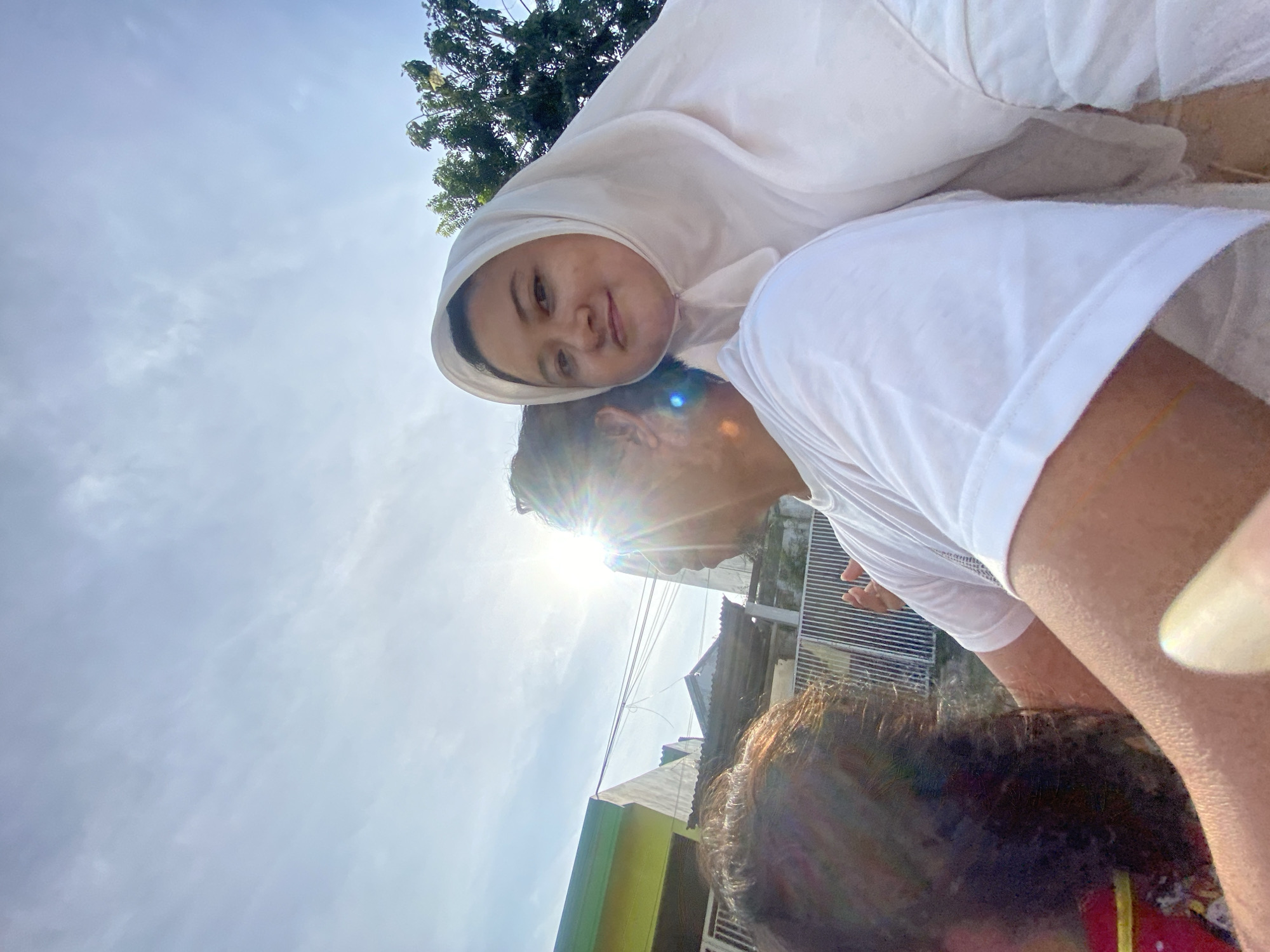 A woman wearing a white headscarf is positioned above a man wearing a white T-shirt. The photo is taken outdoors, possibly in the Gelam, Indonesia based on the prompt. Bright sunlight shines, partly obscuring the man's face. The background includes trees, buildings, and a cloudy sky. The angle suggests the photo may have been taken at an upward angle, or that the people in it are lying or at an angle.