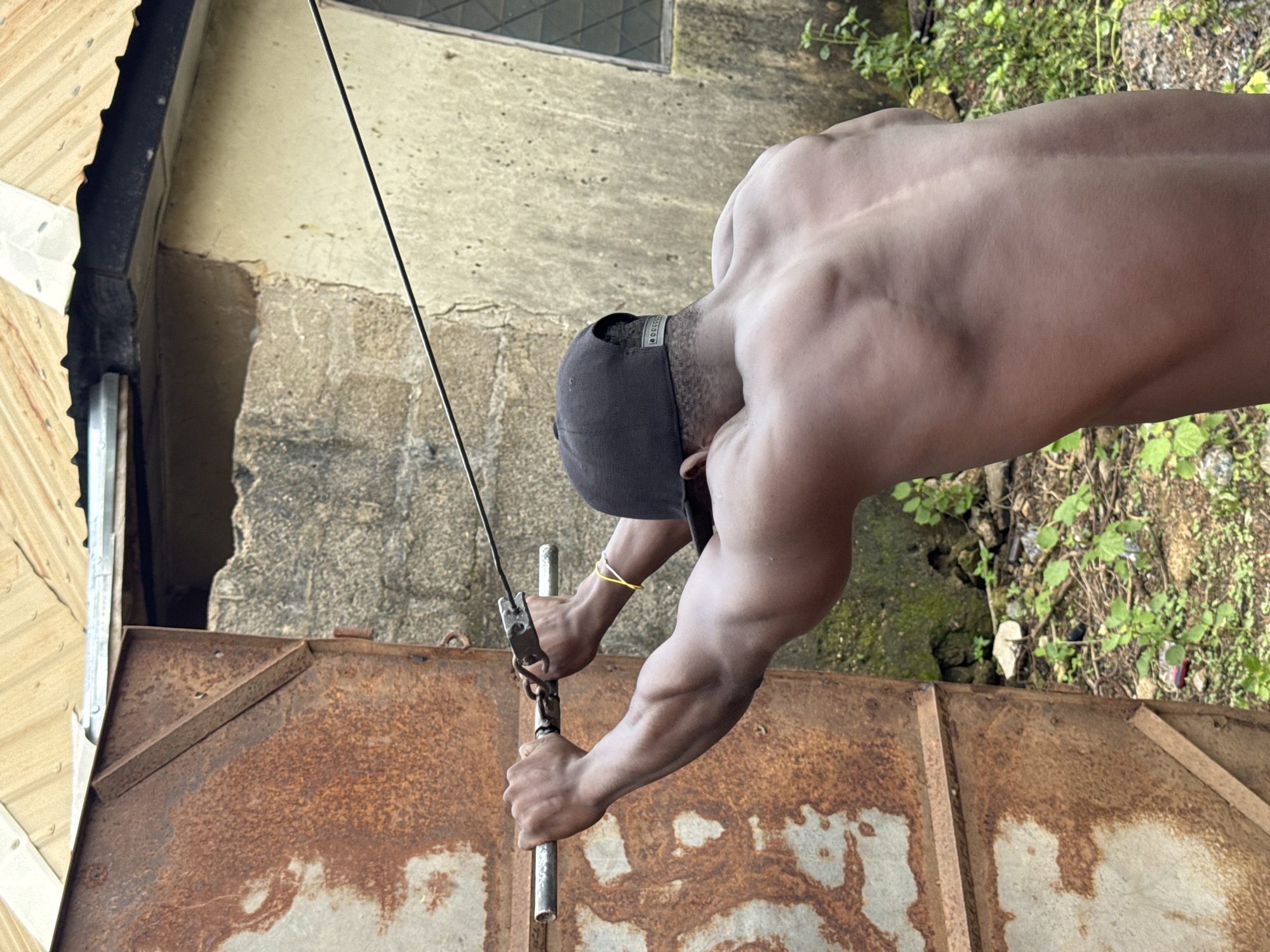 A man, wearing a black baseball cap and a yellow bracelet, is performing a lat pulldown exercise. He is shirtless, revealing his muscular back and arms. The man is gripping a metal bar attached to a cable that runs horizontally across the frame. He is positioned in front of a rusty, weathered metal door, which is partially open. Behind him, a rough, concrete wall and a corrugated metal roof are visible. The ground is covered with dirt, dry leaves, and scattered green vegetation. The scene appears to be outdoors in a rustic, possibly urban or semi-urban, environment.