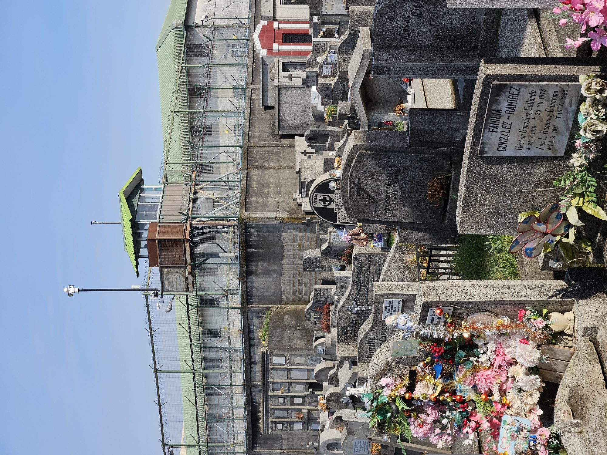 The image shows a cemetery in Osorno, Chile, adjacent to a prison. The scene is divided between the solemn gravestones adorned with flowers and dedications, and the imposing architecture of the prison with its watchtower, wire fences, and security cameras. Details include various tombstones with inscriptions, an abundance of flowers, and the stark contrast between the prison's grey walls and the clear blue sky.
