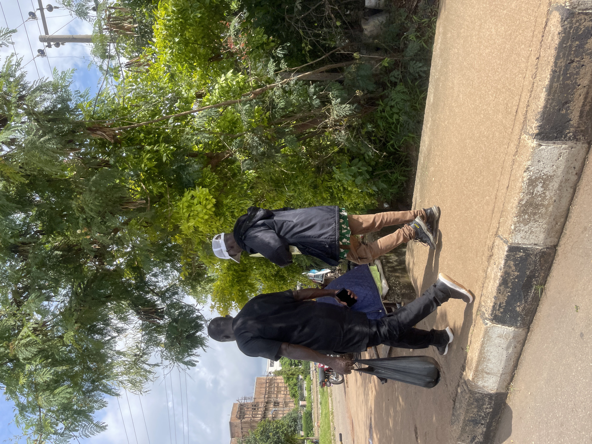 Two men walk along a sidewalk in Barakin Lerawi, Nigeria. The man on the left is wearing a black traditional outfit, black pants, and black sneakers. He is carrying a black plastic bag in his left hand and a black smartphone in his right hand. The man on the right is wearing a black jacket, a white baseball cap, patterned pants, and brown sneakers. Both men are walking away from the camera. The sidewalk is bordered by a concrete curb with black and white markings. Behind the sidewalk is a patch of green vegetation, including trees with lush leaves and some hanging seed pods. In the background, to the left, is a large building under construction, with scaffolding visible. A red motorcycle is parked on the dirt area to the left of the sidewalk. Overhead, power lines are visible against a partly cloudy sky.