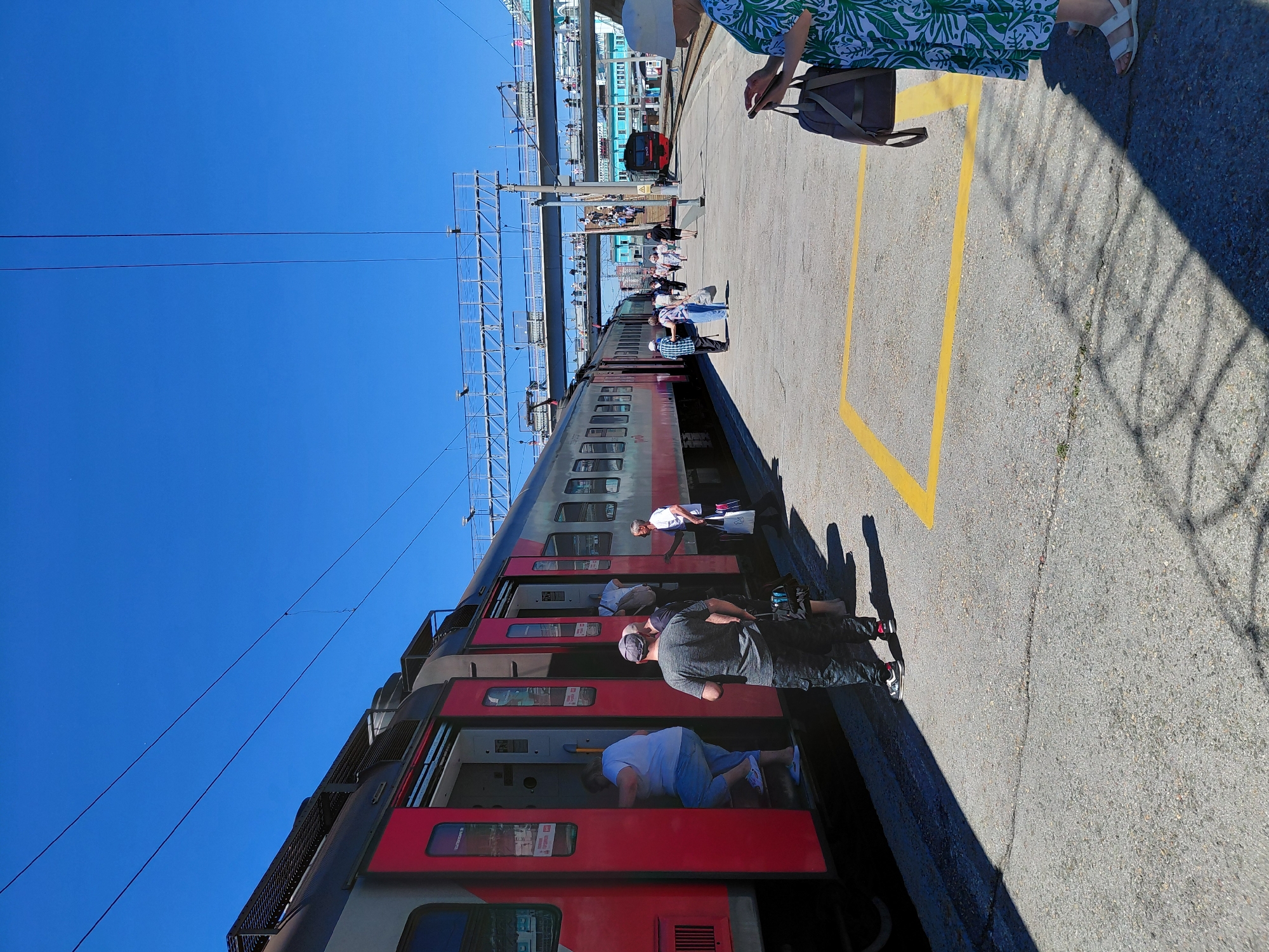 The image depicts a train station scene in Novosibirsk, Russia. A long passenger train, predominantly red and grey, is stopped at the platform. Several people are visible, some boarding or disembarking the train, while others are standing on the platform. The platform is paved with asphalt and marked with yellow lines. In the background, overhead wires for the electric train are visible against a clear blue sky, indicating a sunny day. The time of day appears to be daytime, likely late morning or afternoon, given the bright sunlight and sharp shadows.
Several individuals are dressed in casual clothing. One person is wearing a green and white patterned dress, and another is in a grey t-shirt and dark pants. Some people are carrying bags. The overall atmosphere seems to be that of a typical train station, with people engaged in travel. There are no overt emotional expressions visible, but the activity suggests a sense of movement and transition.
Visible text is limited. Some red signage with white text is present on the train cars, but it is not legible. A yellow line on the platform serves as a safety marking. The scene suggests a functional, operational train station environment.