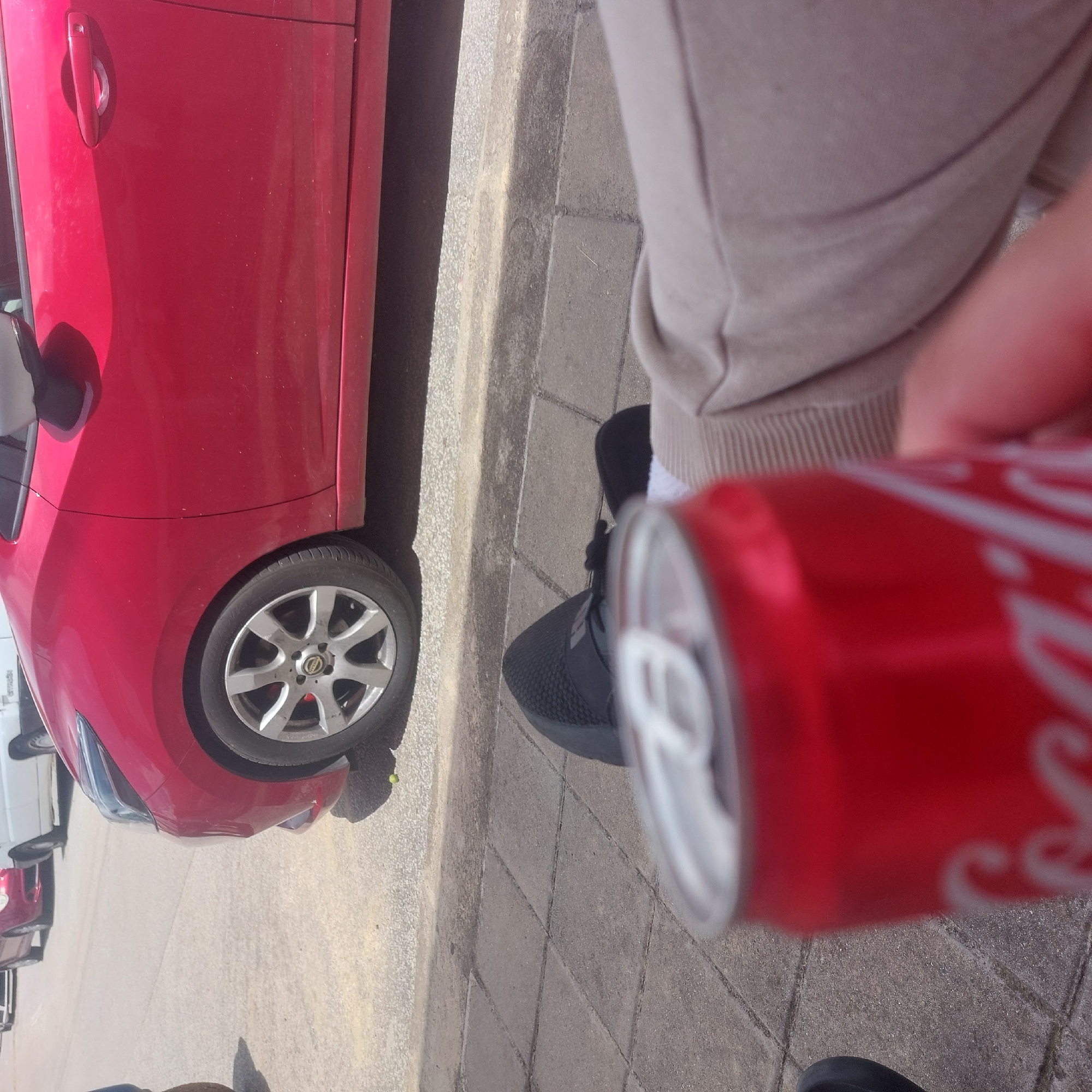 In Ablanitsa, Bulgaria, a person is seated on a sidewalk, holding a Coca-Cola can. The can is red with white lettering and is positioned in the foreground, partially obscuring the person's legs which are clad in beige pants and black athletic shoes. Behind the person, a red car is parked on the street, its front wheel and side visible. The sidewalk is paved with grey square tiles. The background shows a paved street and other vehicles.