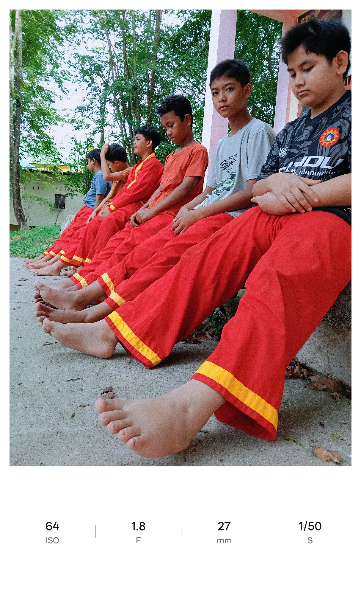 Six boys sit in a row along a concrete ledge, all facing the same direction. The boys are wearing a similar uniform that consists of red pants with yellow bands at the bottom, although they have different shirts on. They are all barefoot and their feet are visible, lined up on the concrete in front of the ledge. The background consists of greenery, trees, and a building with a green roof. The scene appears to be a candid shot with the subjects positioned in front of the camera with limited interaction. The image includes metadata indicating an ISO of 64, an aperture of f/1.8, a focal length of 27mm, and a shutter speed of 1/50s. The image is identified as being in Lhokseumawe, Indonesia.