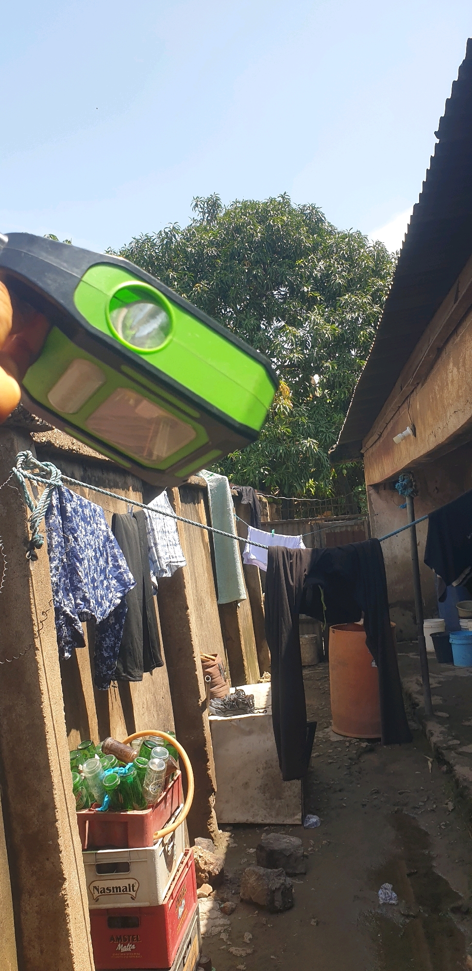 The image captures a sunlit outdoor scene in Sabon Tasha, Nigeria, likely a residential or backyard area. A hand is holding a bright green and black object, possibly a portable light or speaker, positioned in the foreground and obscuring part of the view. In the background, a large, leafy tree dominates the center, with a corrugated metal roof of a building visible on the right. Clothes are hung on a clothesline strung between concrete posts, including patterned fabric, black trousers, a white garment, and a light blue towel. Below the clothesline, a red crate filled with green glass bottles and a yellow hose lies on top of a white crate labeled "Nasimalt" and a red crate labeled "AMSTEL MALTA". The ground appears to be a mix of mud and scattered debris, with a large orange barrel visible on the right side of the image. The sky is clear and blue.